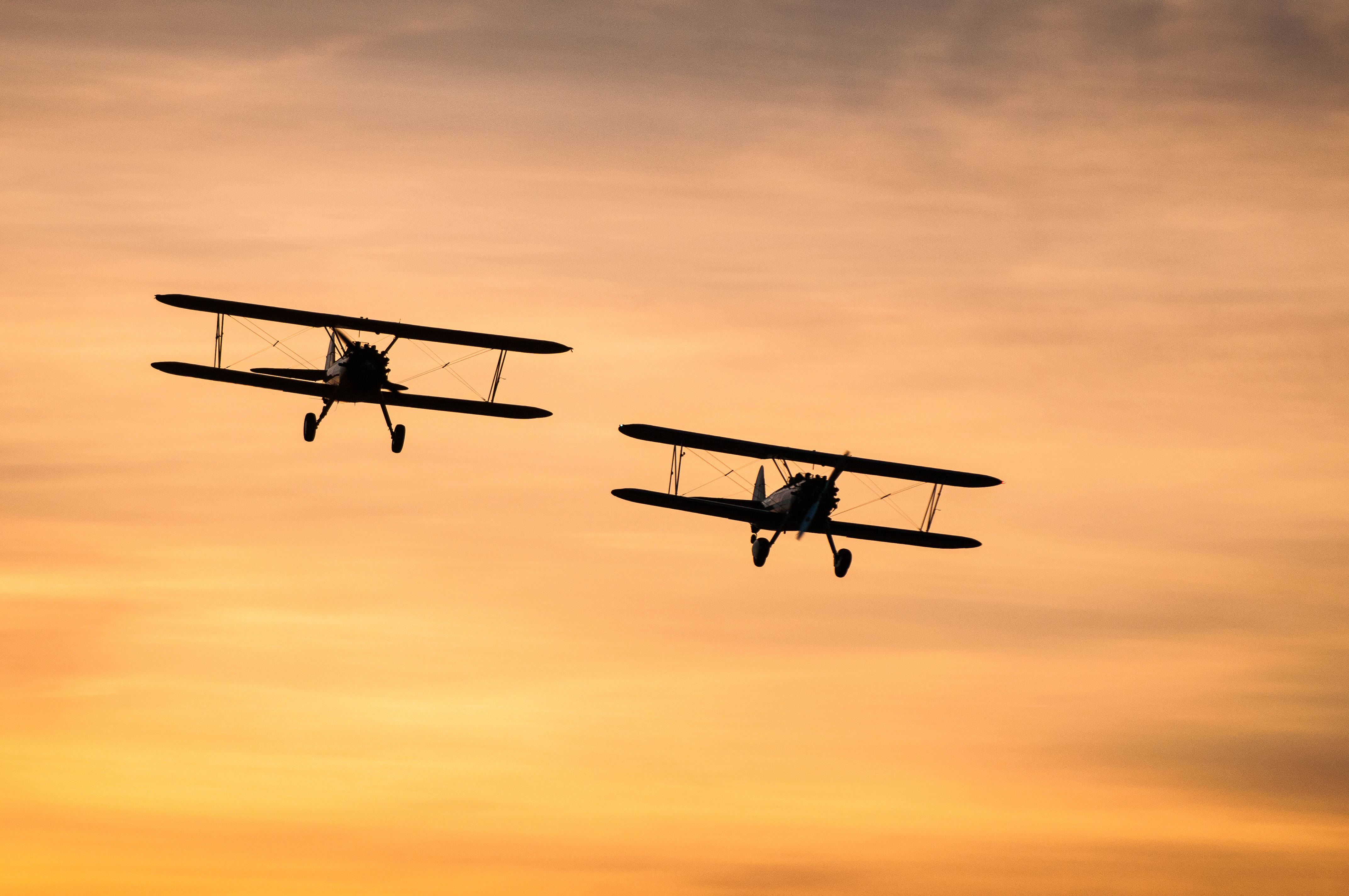 夕陽天空中的小型飛機 壁紙 — 兩架復古雙翼機在火紅橙黃的夕陽天空背景下翱翔，呈現剪影。