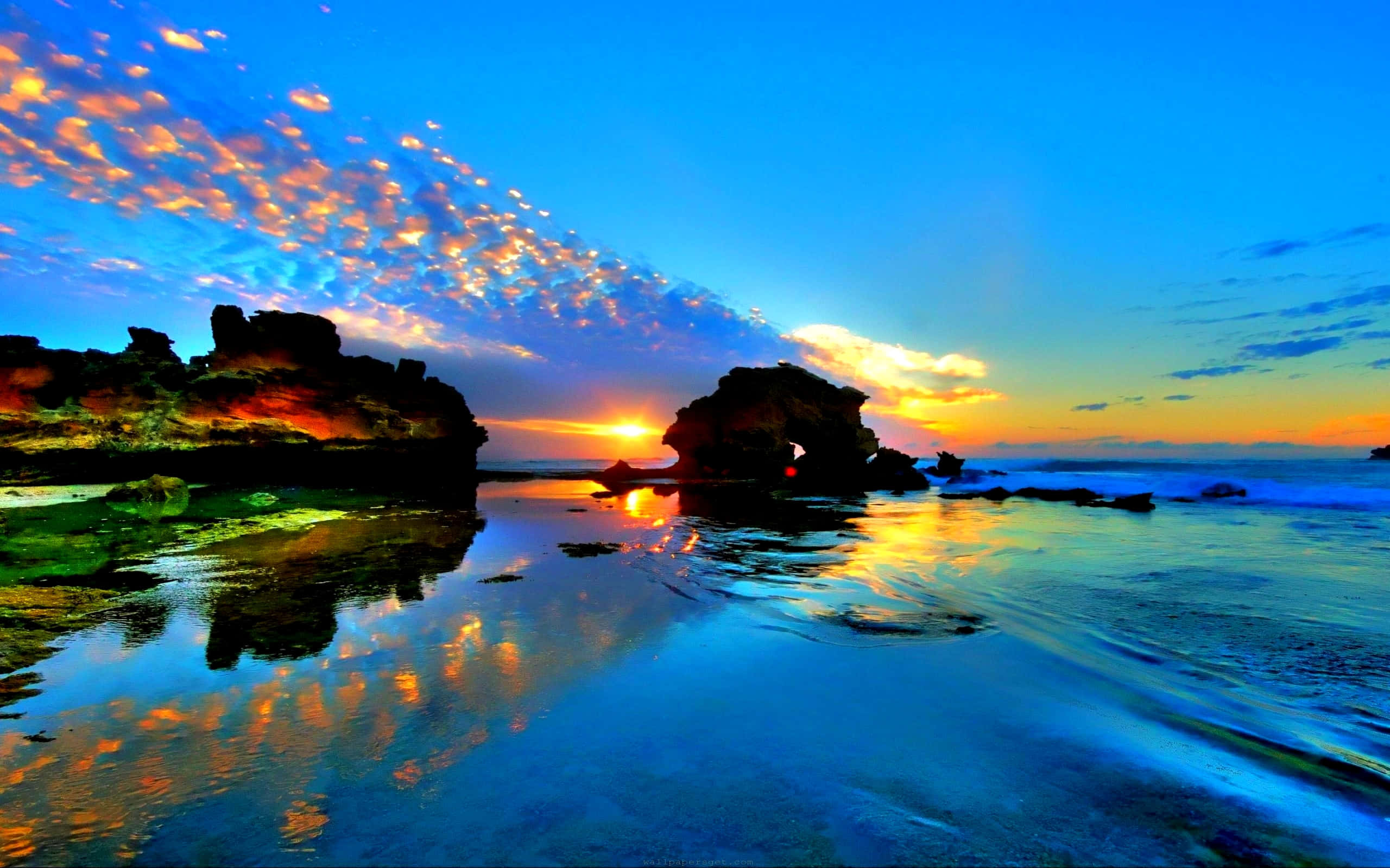 Beautiful Beach 桌布 壁紙 — 夕陽將天空和海洋染成火熱的橙色和深藍色，海浪拍打著壯觀的岩石 formations。