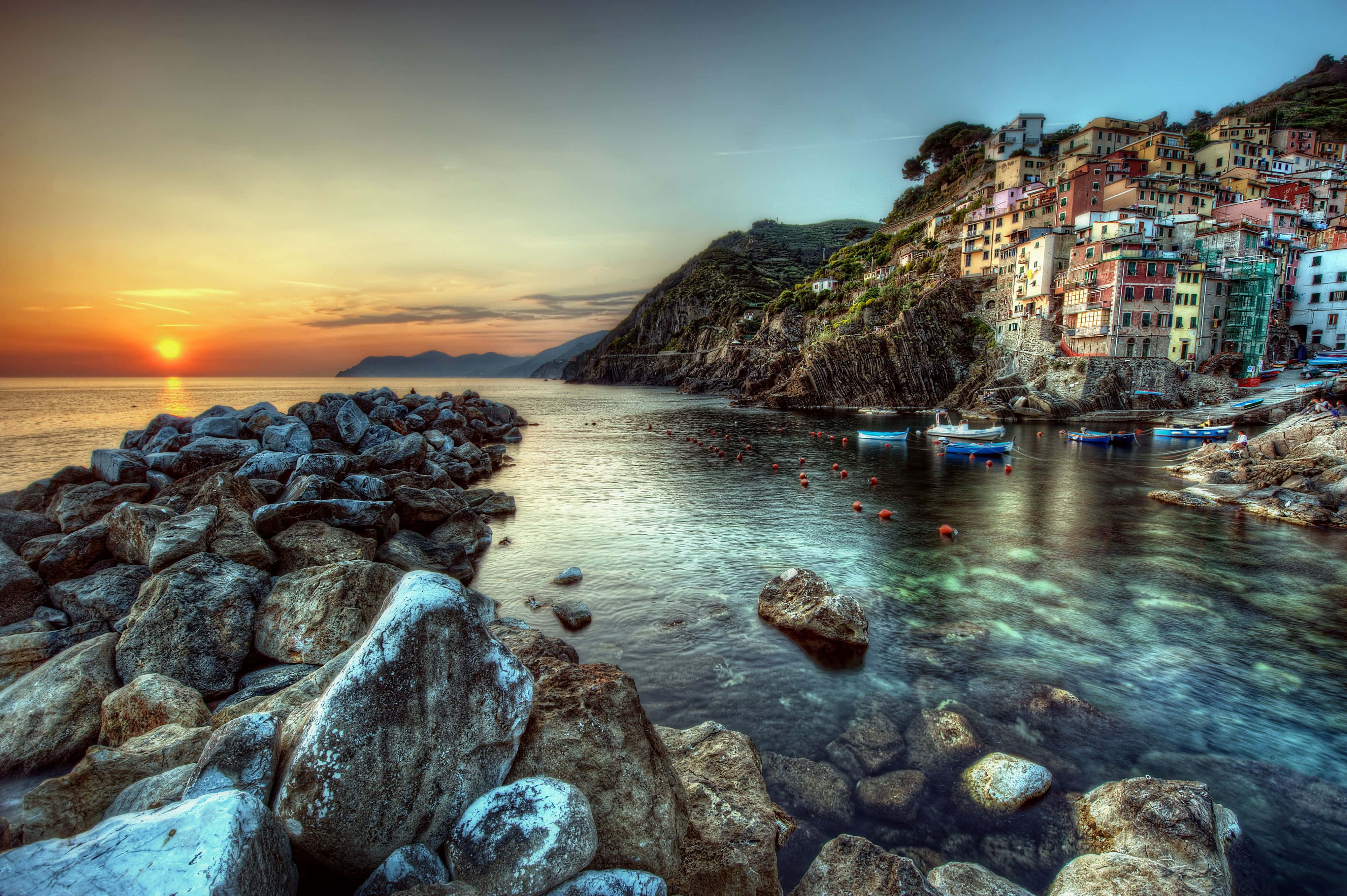 4K Hdr 桌布 壁紙 — 夕陽將天空染成橙色，色彩繽紛的懸崖房屋俯瞰著點綴著船隻和岩石海岸的平靜水域。