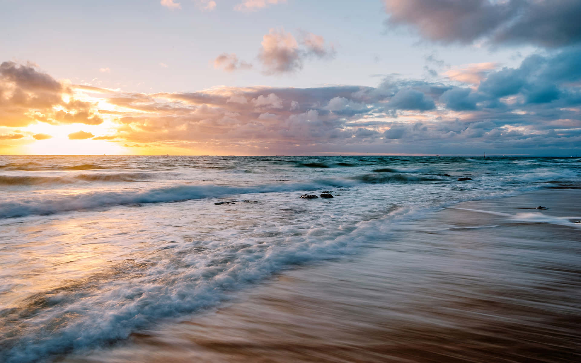 Beach Waves 桌布 壁紙 — 夕陽將天空染成溫暖的金色和柔和的粉紅色，海浪在戲劇性的雲層下拍打著沙灘。