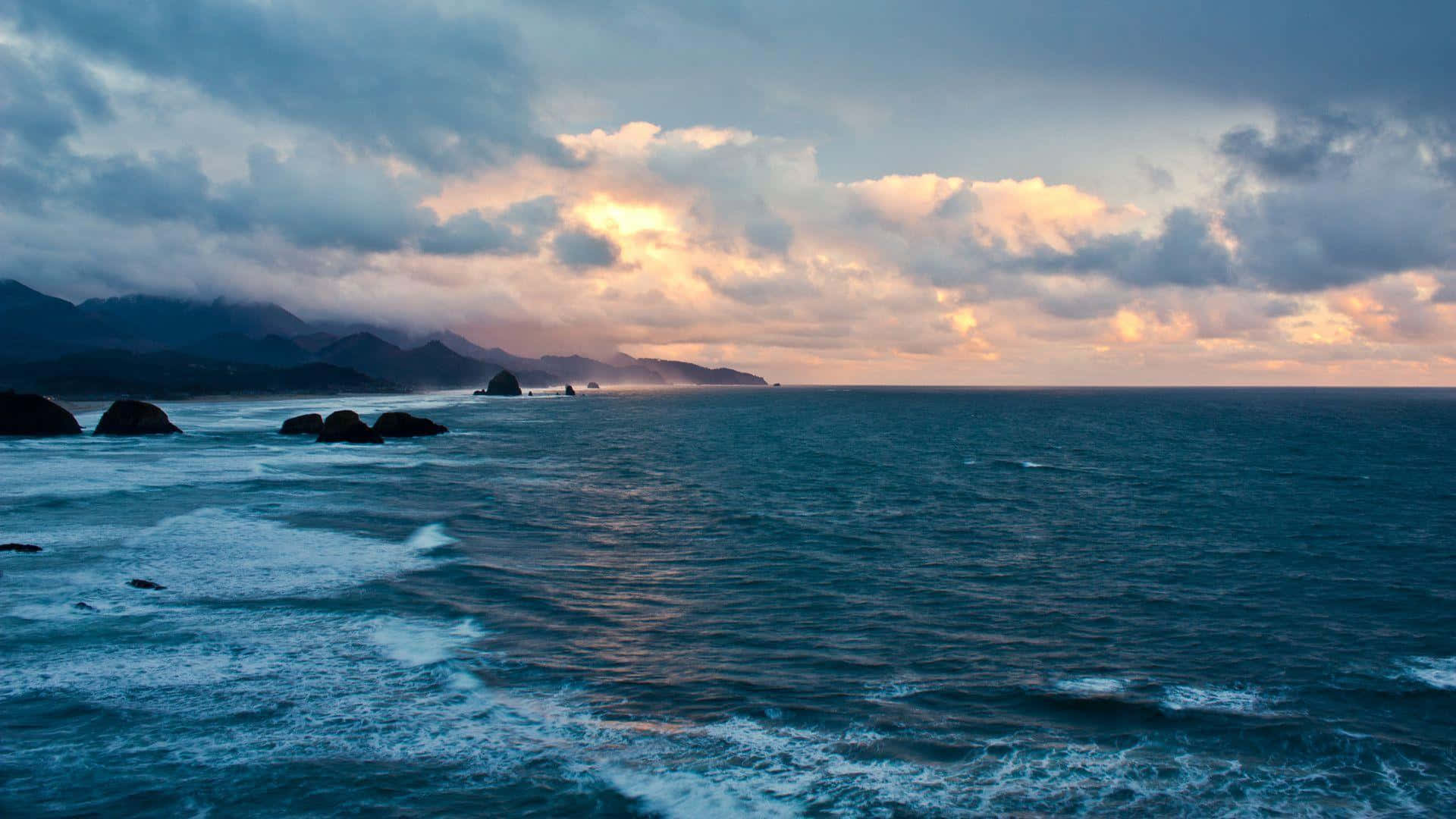 Beautiful Ocean 桌布 壁紙 — 夕陽將天空染成溫暖的金色和柔和的粉紅色，下方是波濤洶湧的海洋，遠處有烏雲和山脈。