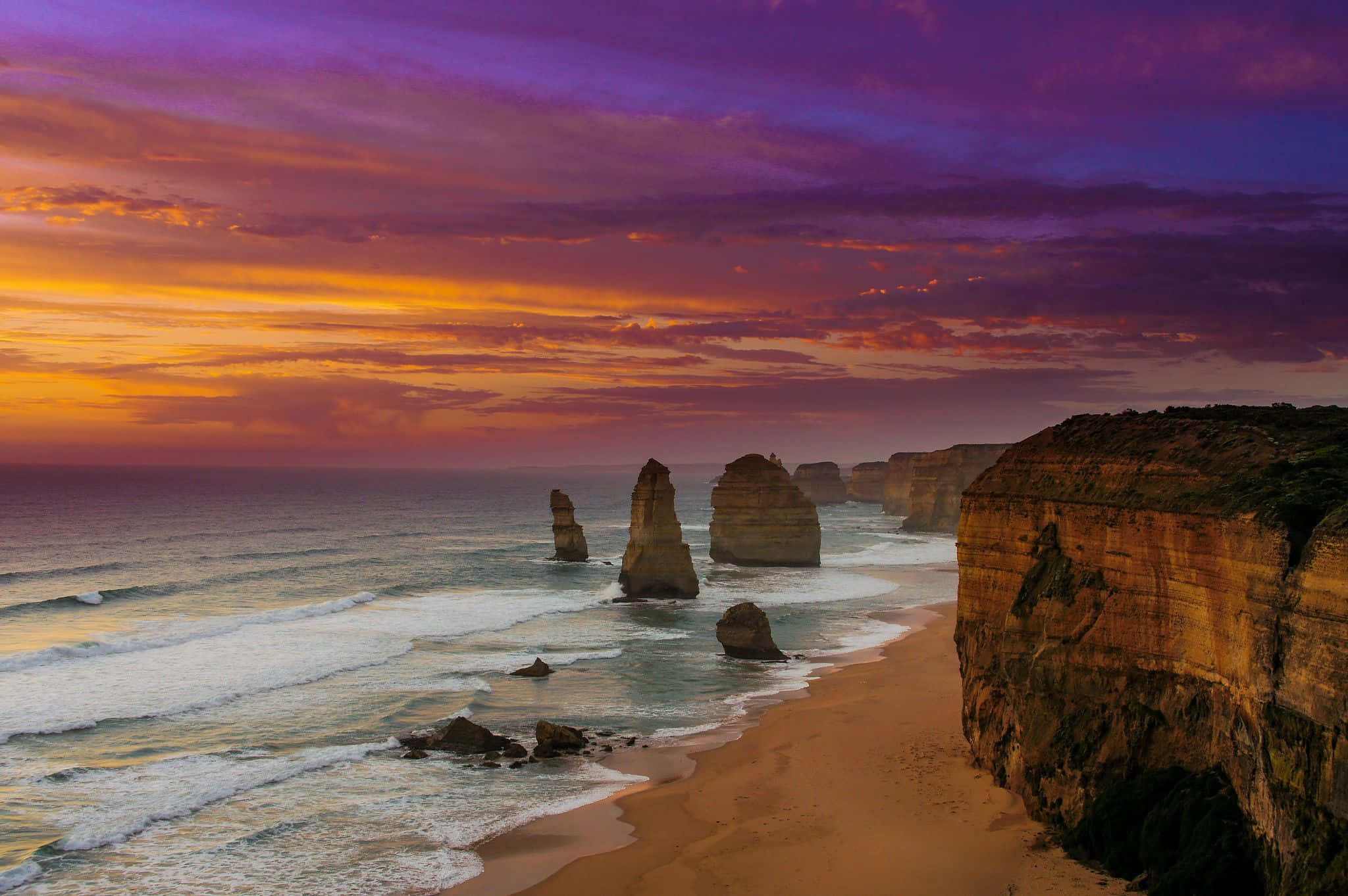 Australia Beach 桌布 壁紙 — 夕陽將天空染成火橙色和深紫色，映襯著從海洋中升起的標誌性十二門徒石灰岩柱。