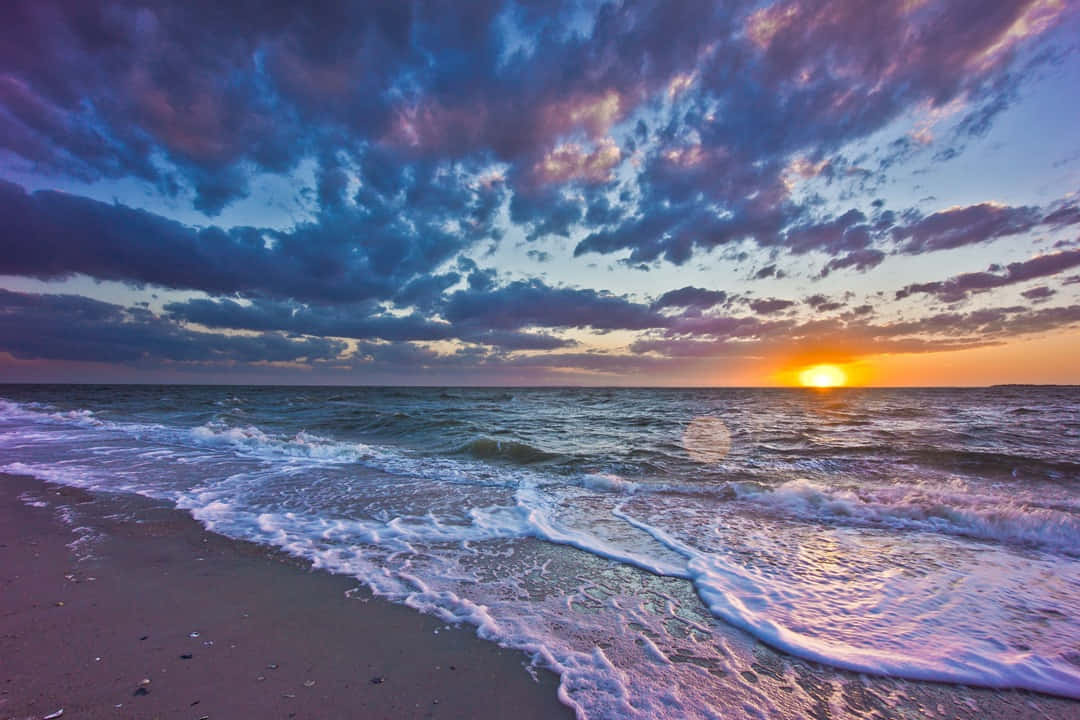 Sunset Wave 桌布 壁紙 — 夕陽將天空染成火橙色和深紫色，海浪在戲劇性的雲層下拍打沙灘。