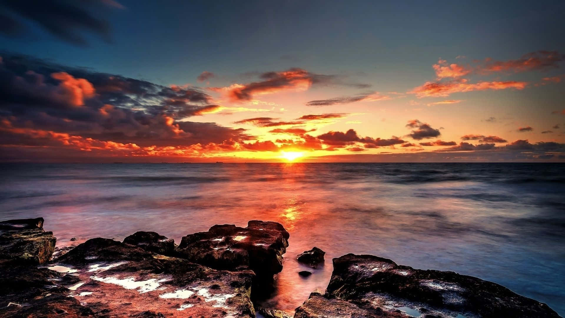 4K Hdr 桌布 壁紙 — 夕陽將天空染成火橙色和深藍色，海浪輕輕拍打岸邊的黑色岩石。