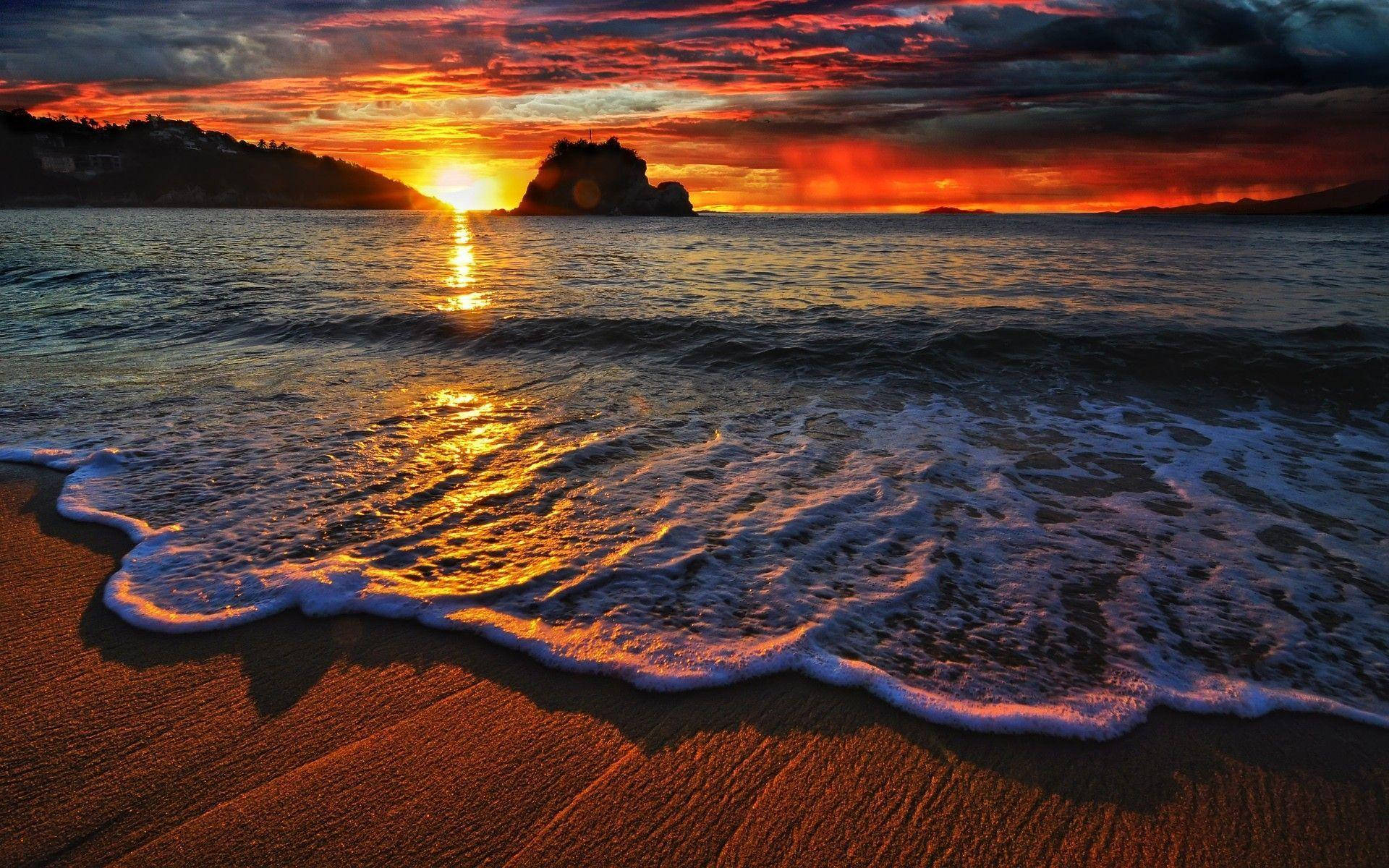 日落 桌布 壁紙 — 夕陽將天空染成火橙色和紅色，海浪拍打沙灘，遠處有岩石島的輪廓。