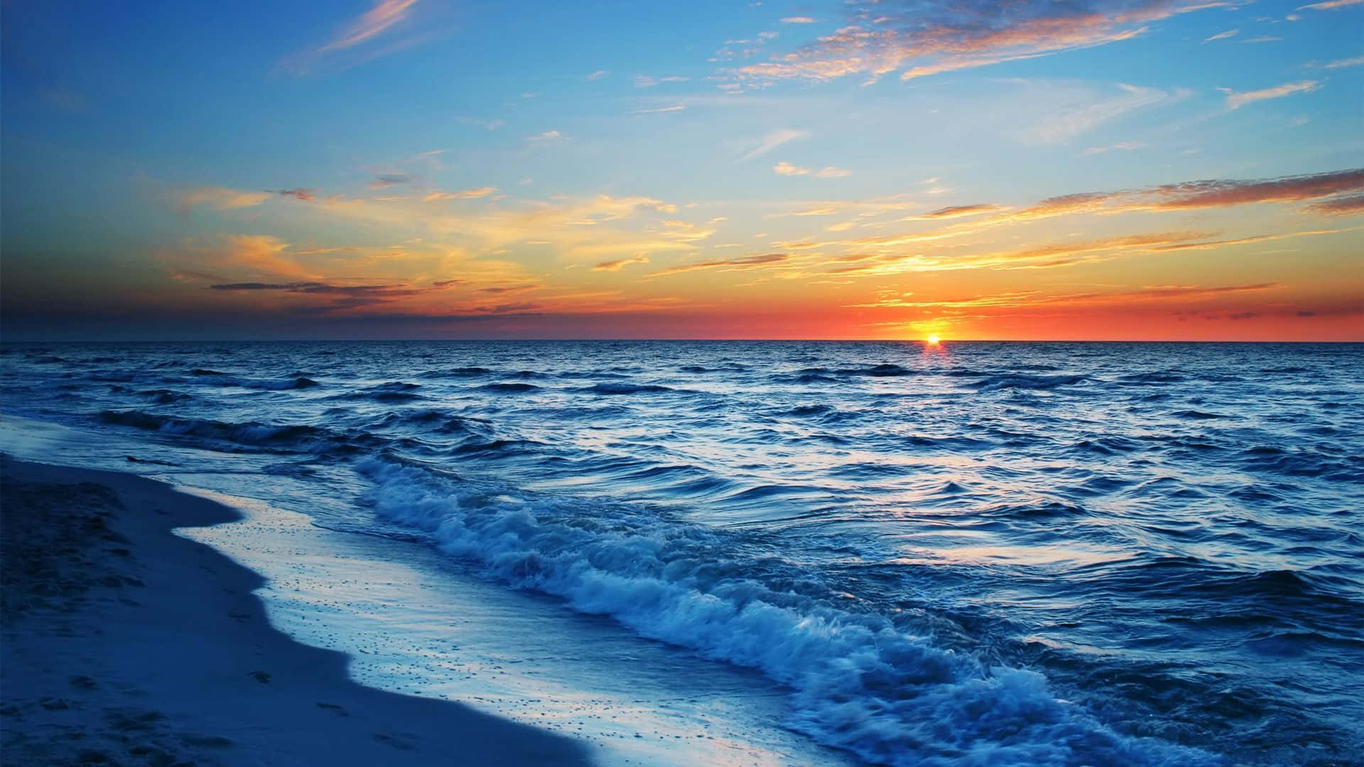 Sunset Ocean 桌布 壁紙 — 夕陽將天空染成火熱的橙色和粉紅色，海浪在廣闊的藍色地平線下拍打著沙灘。