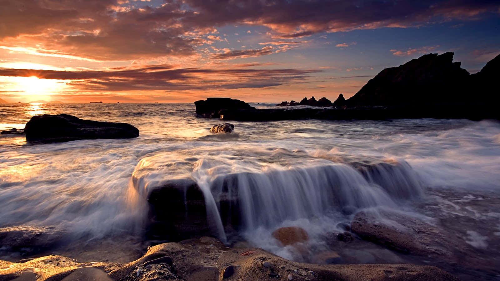 3d Ocean 桌布 壁紙 — 夕陽將天空染成火熱的橙色和紫色，海浪在寧靜的海岸場景中翻滾過岩石。