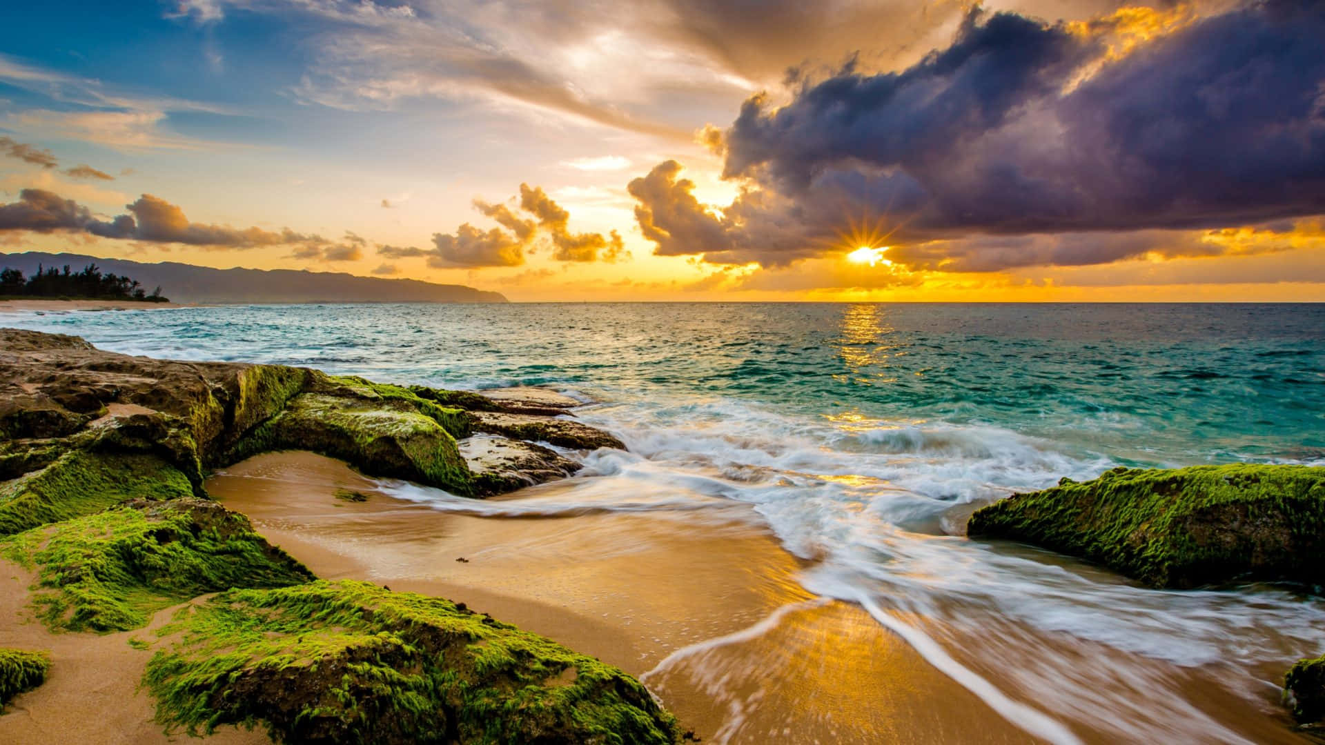 Sunset Ocean 桌布 壁紙 — 夕陽將天空染成火熱的橙色和紫色，海浪拍打著熱帶海灘上長滿苔蘚的岩石。