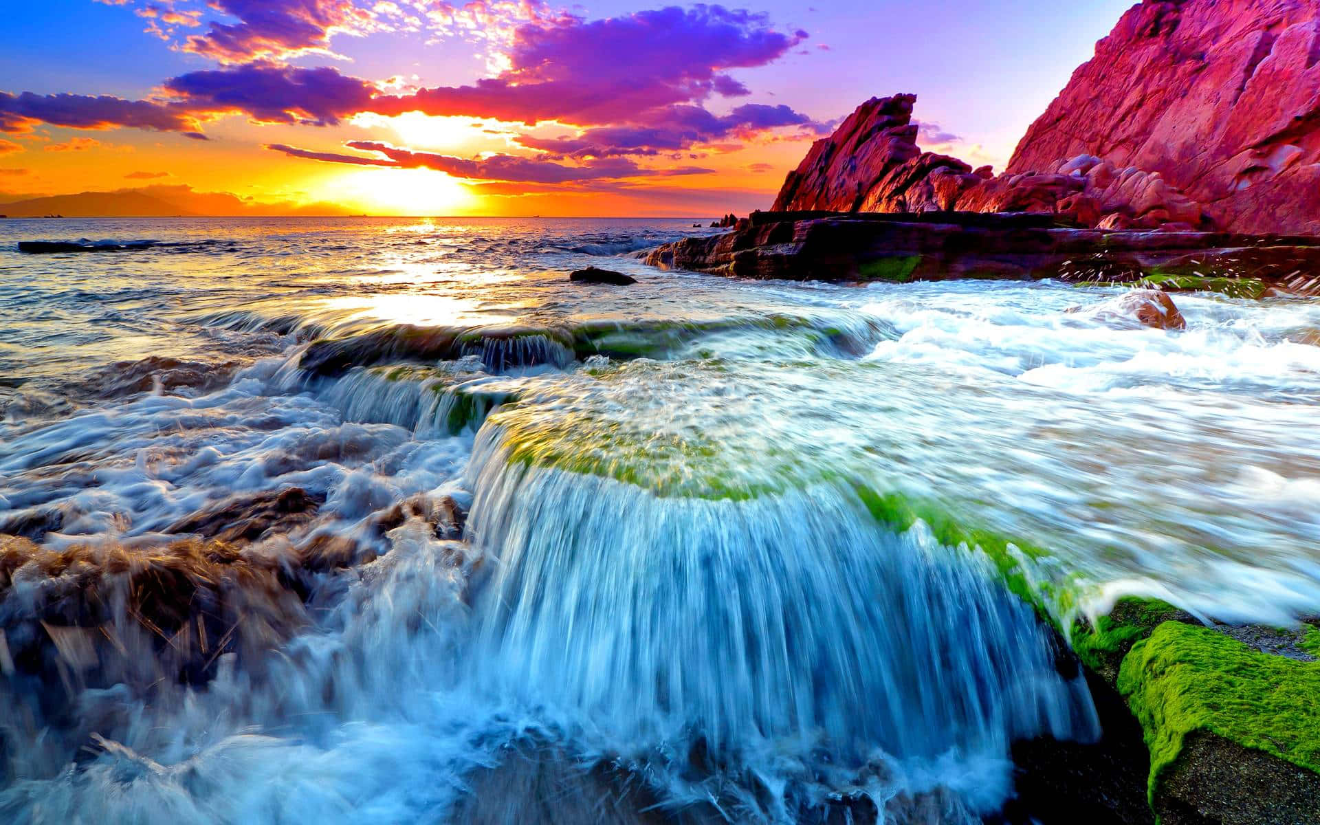 3d Ocean 桌布 壁紙 — 夕陽將天空染成火熱的橙色和紫色，海浪拍打長滿苔蘚的岩石，形成碧綠色的瀑布。