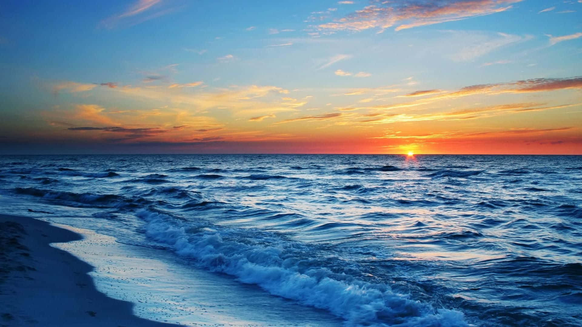 海灘日落 桌布 壁紙 — 夕陽將天空染成火熱的橙色和藍色，海浪拍打著沙灘。