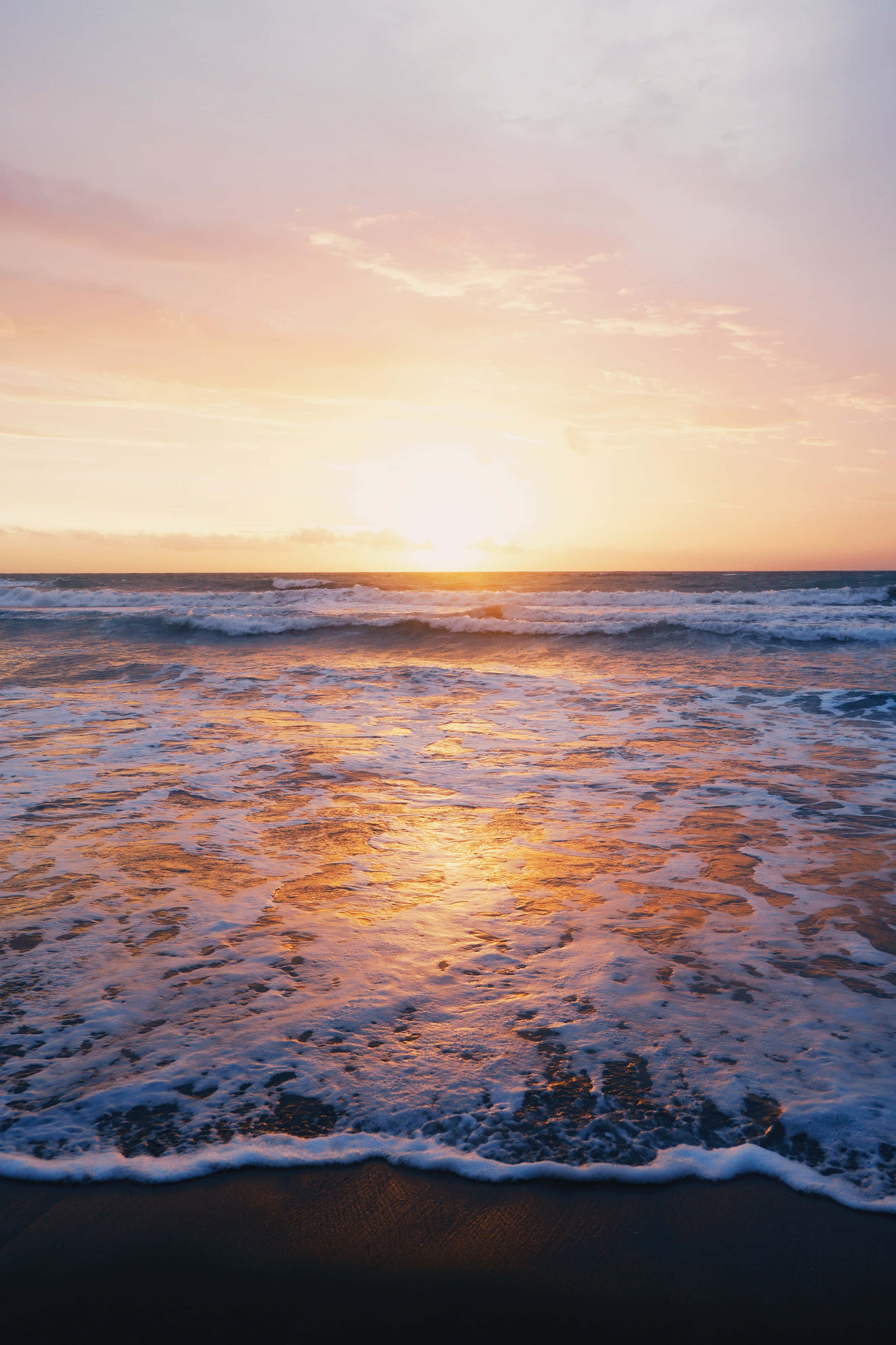 Beach Photography 桌布 壁紙 — 夕陽將海洋染成溫暖的金色和粉紅色，海浪拍打著深色的沙灘。