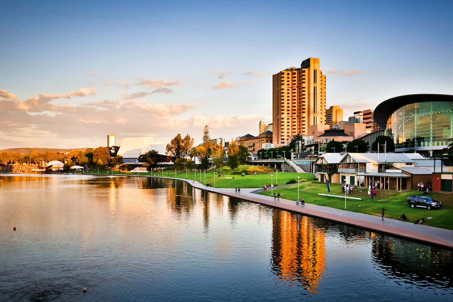 Adelaide 桌布 壁紙 — 夕陽將現代城市天際線和寧靜的湖泊沐浴在金色光芒中，人們沿著海濱小徑漫步。