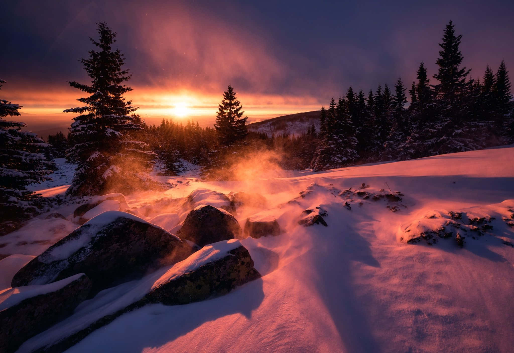 Winter Sun 桌布 壁紙 — 夕陽將積雪的山脈和松樹染成火熱的橙色，蒸汽從積雪覆蓋的岩石上升起。