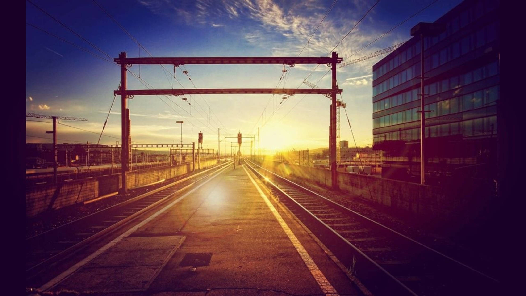 日出 桌布 壁紙 — 夕陽將空無一人的火車軌道沐浴在金色光芒中，軌道延伸向一座現代玻璃建築，上方是充滿活力的藍天。