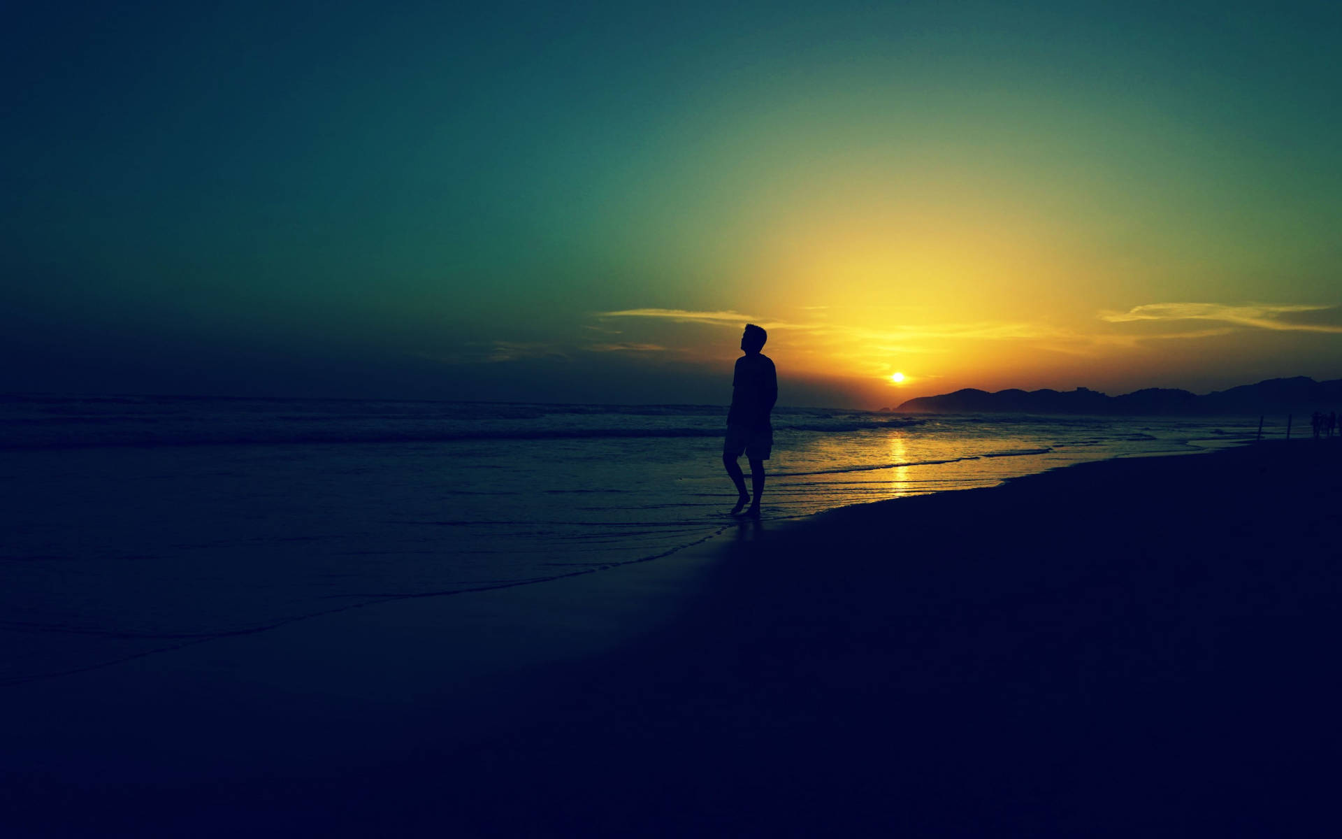 夕陽海岸獨行 壁紙 — 一個孤獨的身影站在沙灘上，看著夕陽落在遠山之上，在水面上灑下金色的光芒。