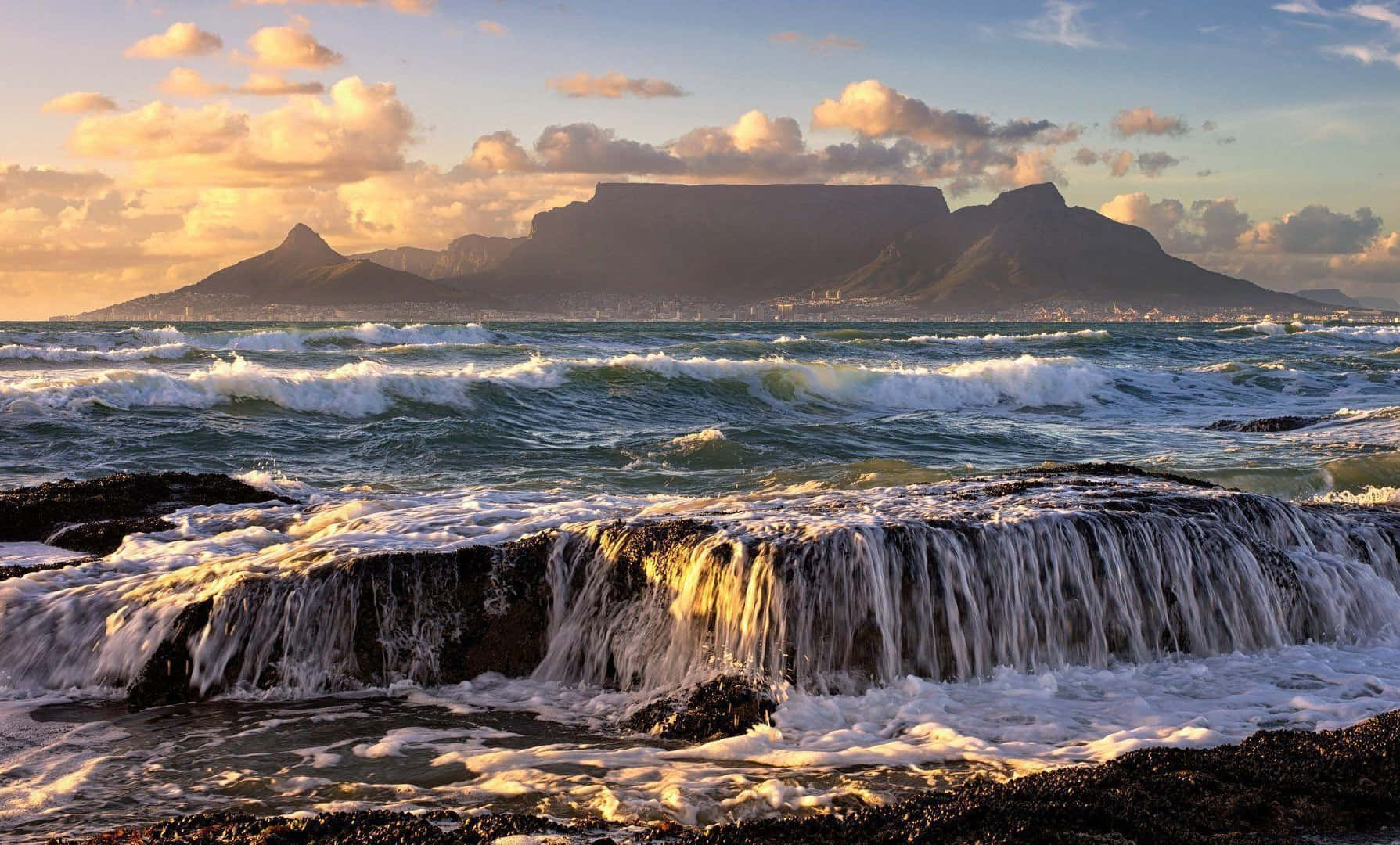 South Africa 桌布 壁紙 — 夕陽海浪拍打黑色岩石，桌山在金色雲層天空的襯托下呈現剪影。