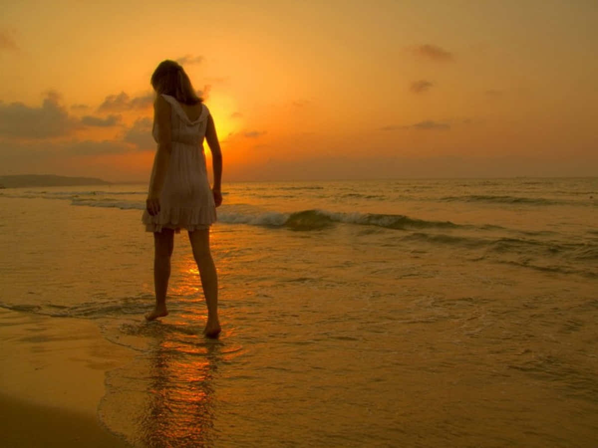 夕陽海灘獨坐憂鬱女子 壁紙 — 一位身穿白色連衣裙的女子在日落時赤腳站在沙灘上，金色的海浪輕拍她的雙腳。