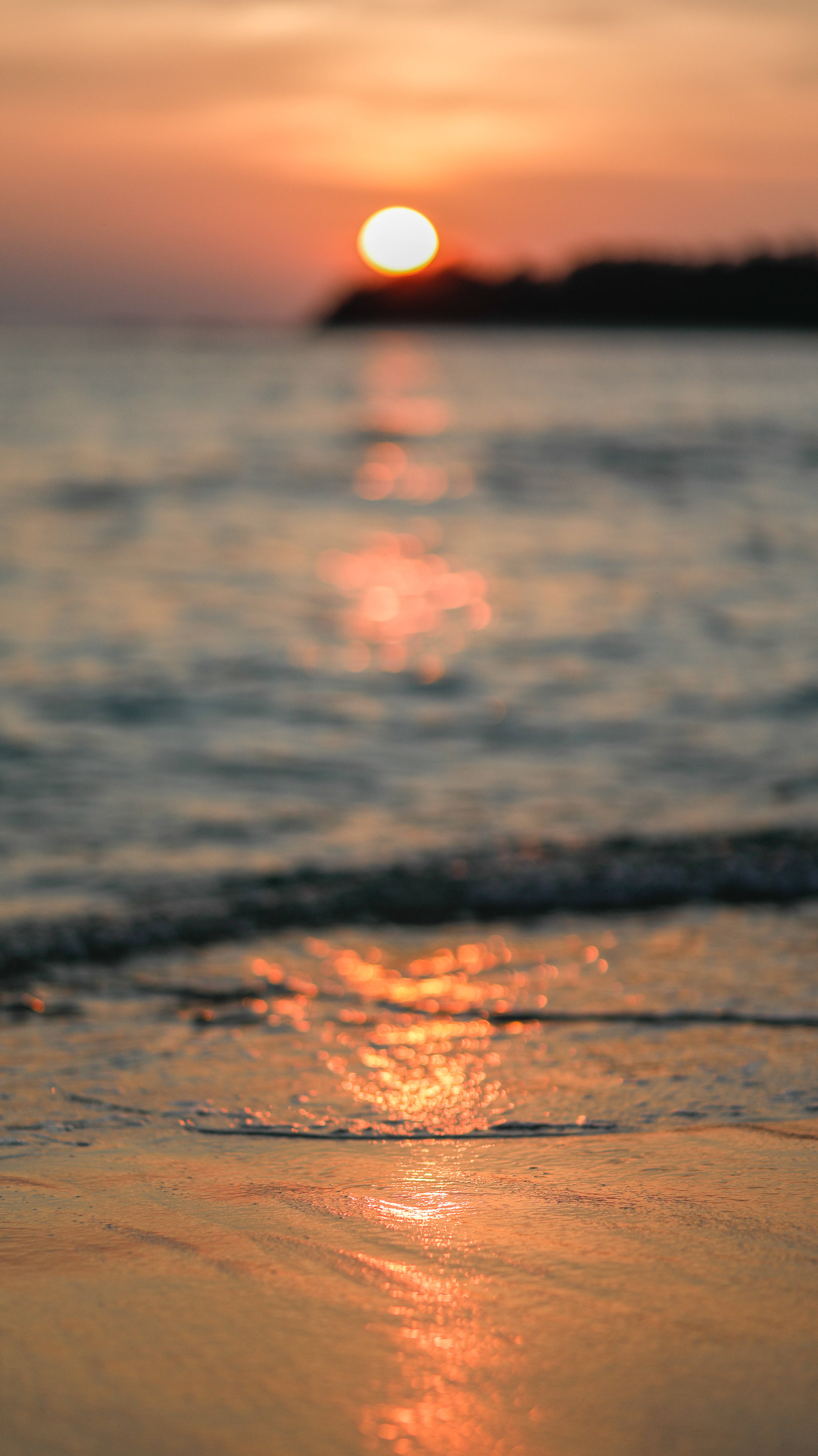 日落 桌布 壁紙 — 夕陽溫暖地照耀著海浪拍打沙灘，遠處有陸地輪廓。