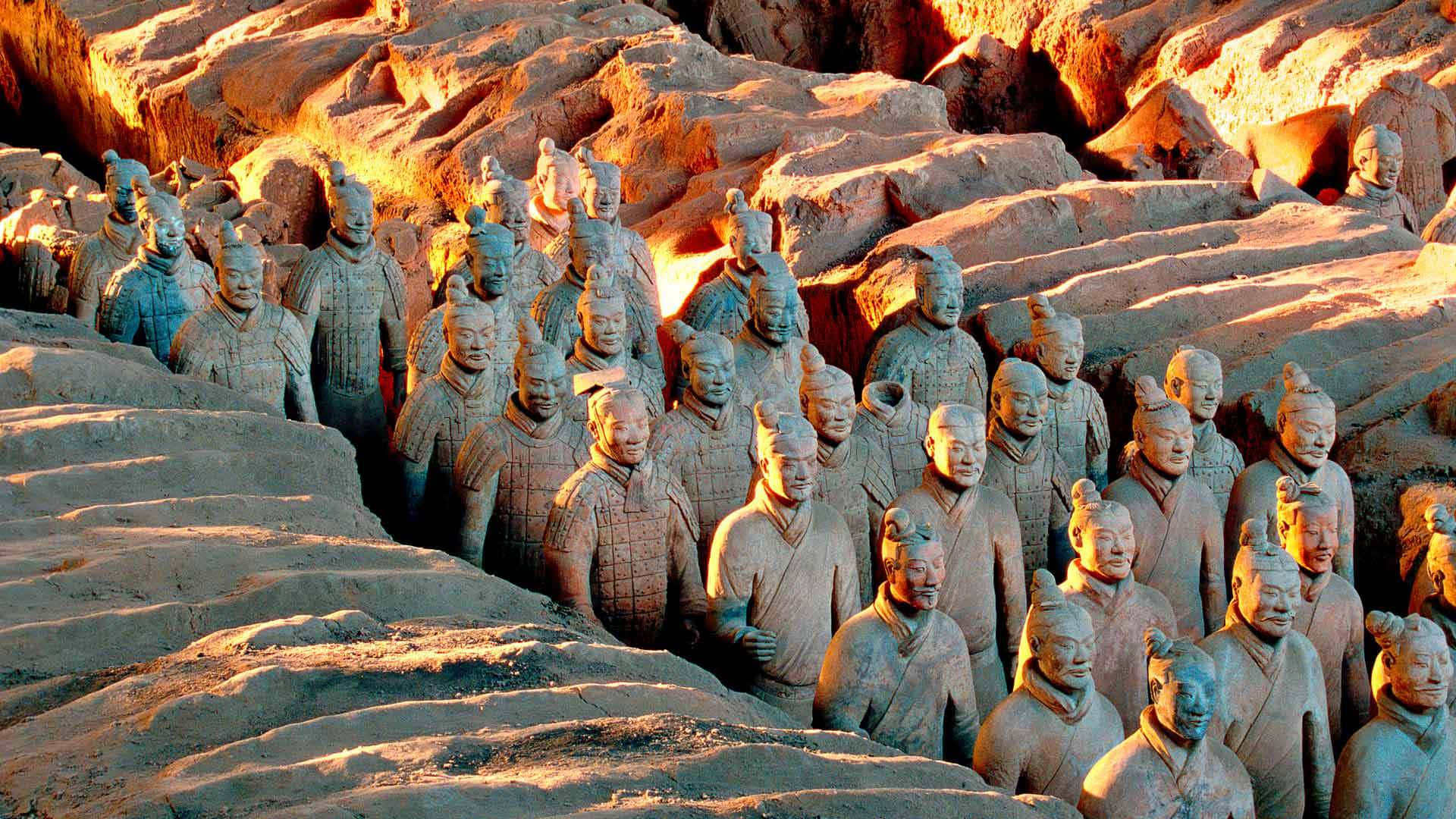 夕陽灑落的秦俑雕塑 壁紙 — 一排排古老的兵馬俑從泥土戰壕中浮現，沐浴在溫暖的金色夕陽光中。