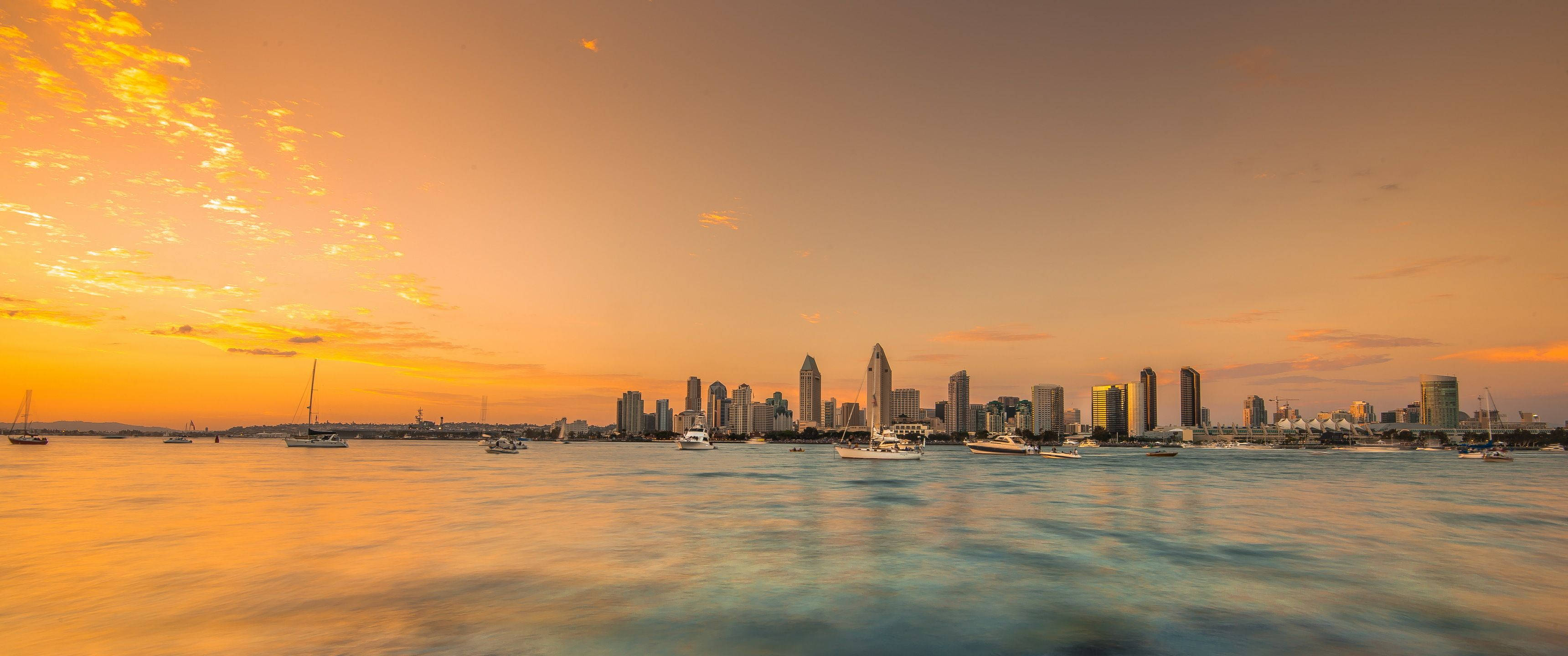 View 桌布 壁紙 — 夕陽燦爛，將聖地牙哥的天際線染成金色，船隻在下方平靜的水面上滑行。