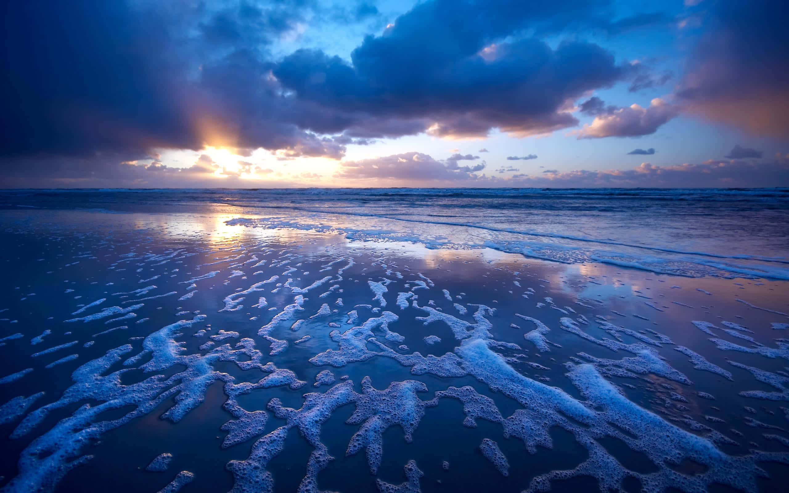 Beautiful Ocean 桌布 壁紙 — 夕陽的光線穿過暴風雲，將金色光芒投射在濕潤沙灘上翻滾的泡沫海浪上。