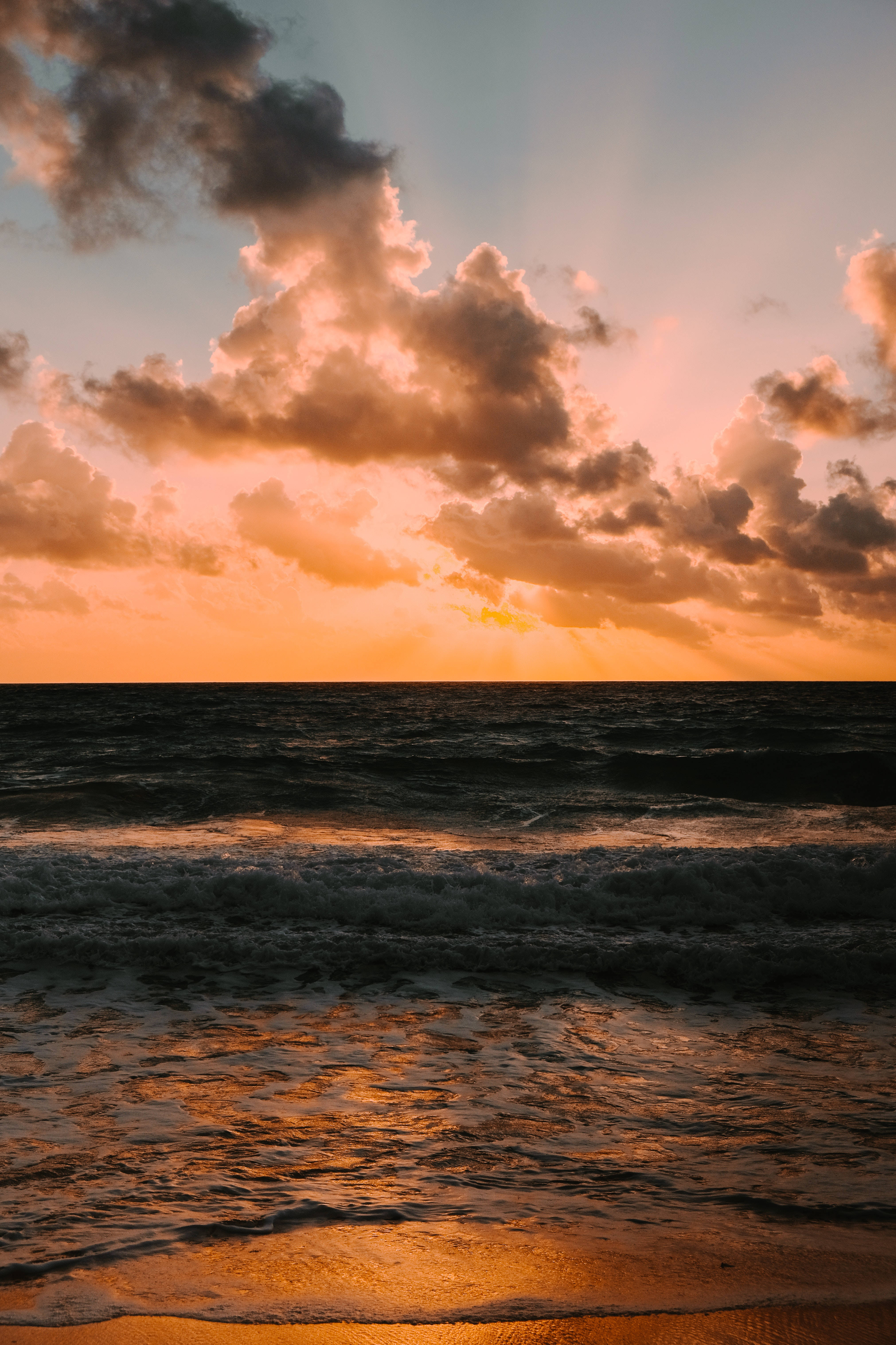 日落 桌布 壁紙 — 夕陽穿透戲劇性的雲層，照耀在波濤洶湧的海洋上，在濕沙上反射出熾熱的橙色和粉紅色調。