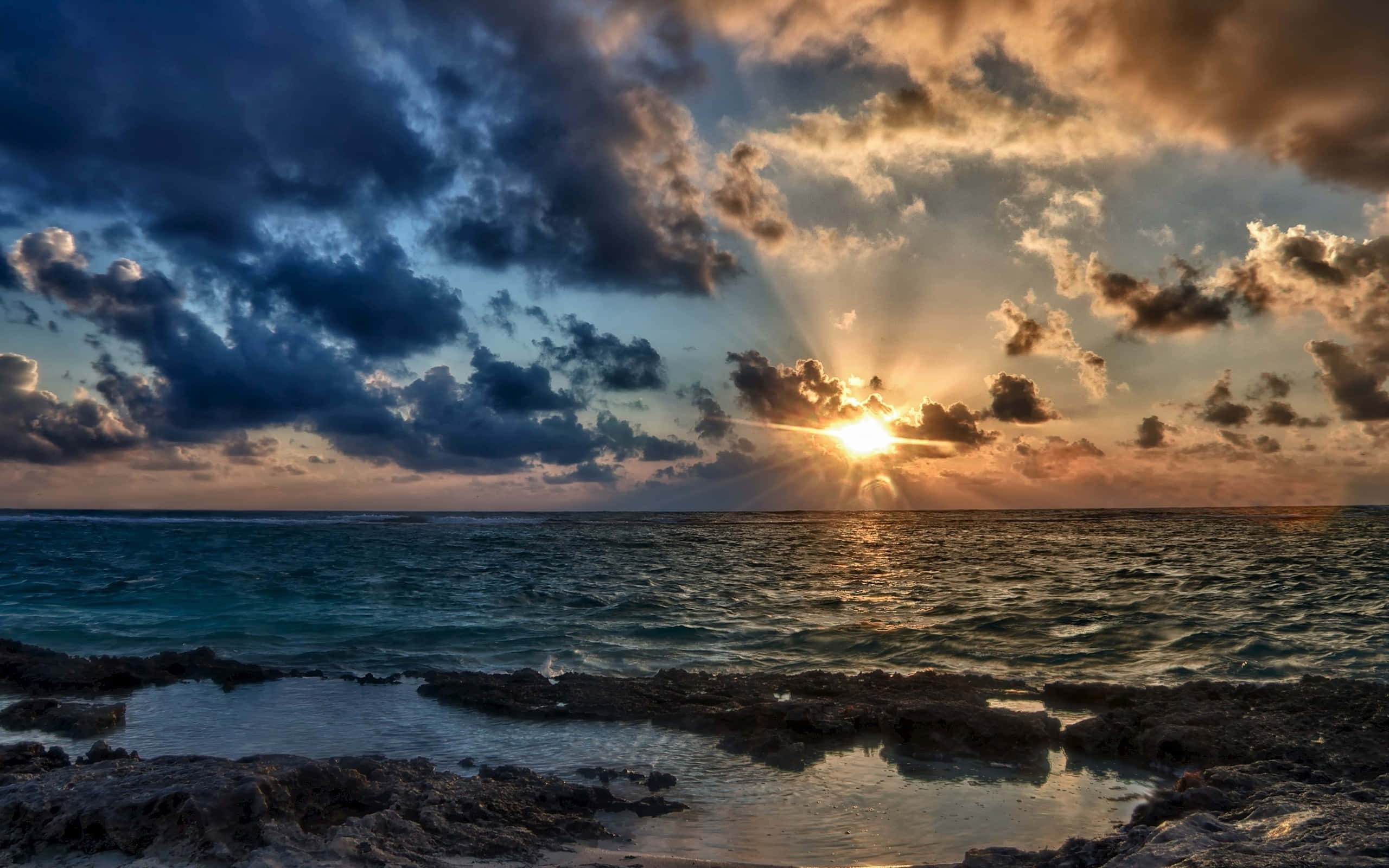 Sunset Ocean 桌布 壁紙 — 夕陽穿透戲劇性的雲層，照耀在碧綠海洋與岩石海岸交匯處。