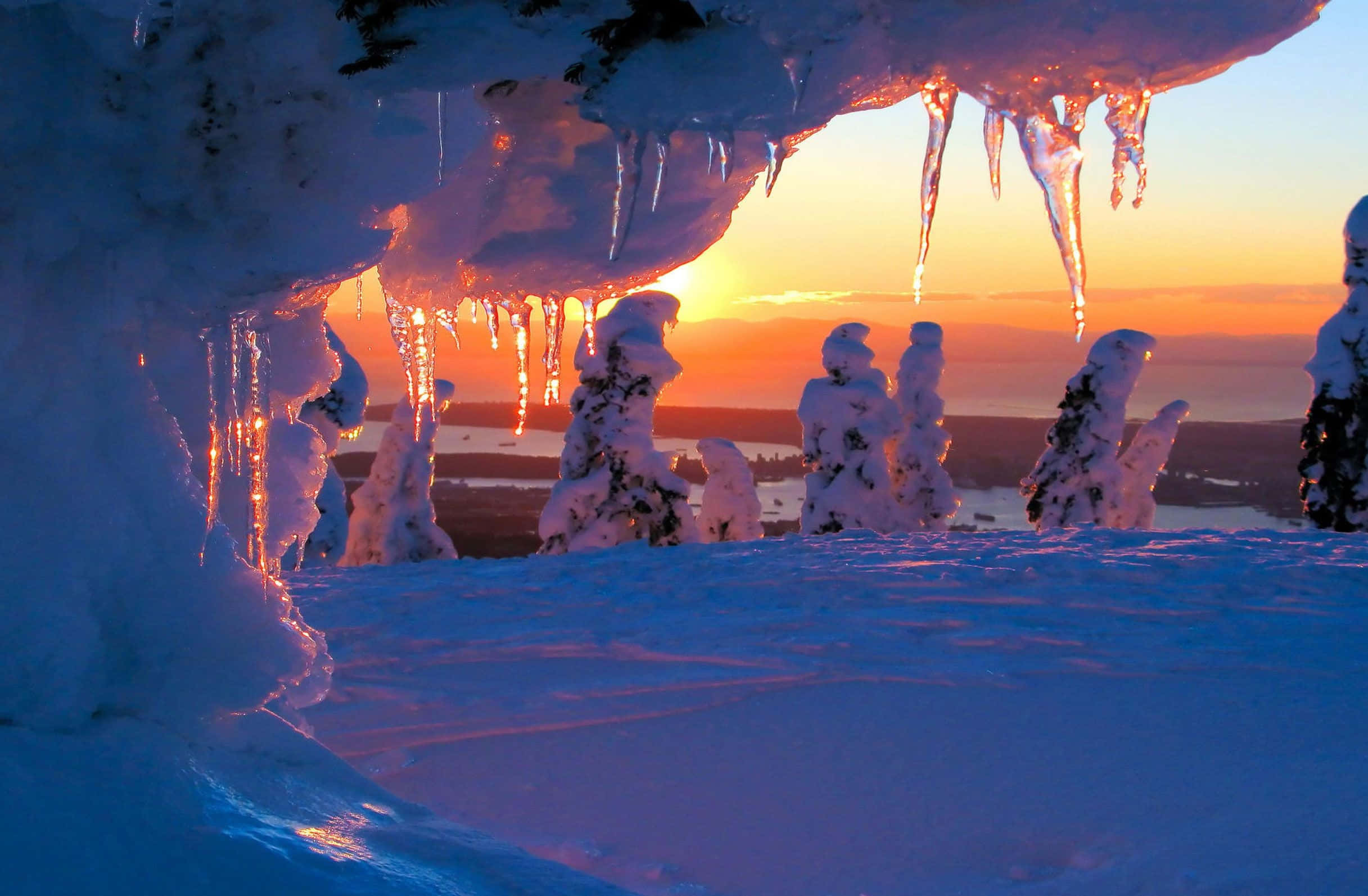 Winter Sunset 桌布 壁紙 — 夕陽穿過冰簷閃耀，在冬日景觀中將溫暖光線投射在積雪覆蓋的樹木和冰柱上。