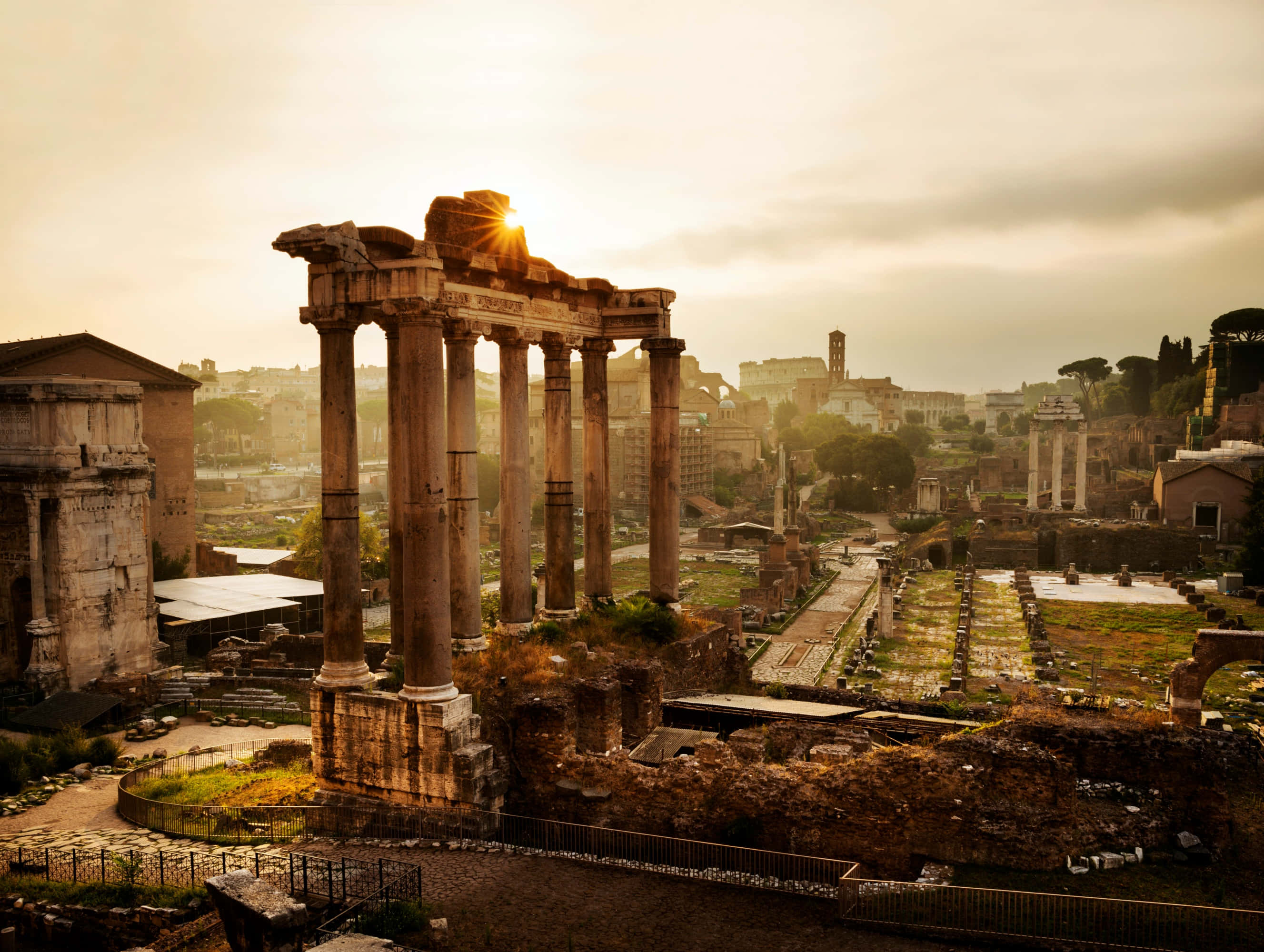 Roman 桌布 壁紙 — 夕陽穿過古羅馬柱廊，在風化的廢墟和遠處的城市景觀上投下長長的影子。