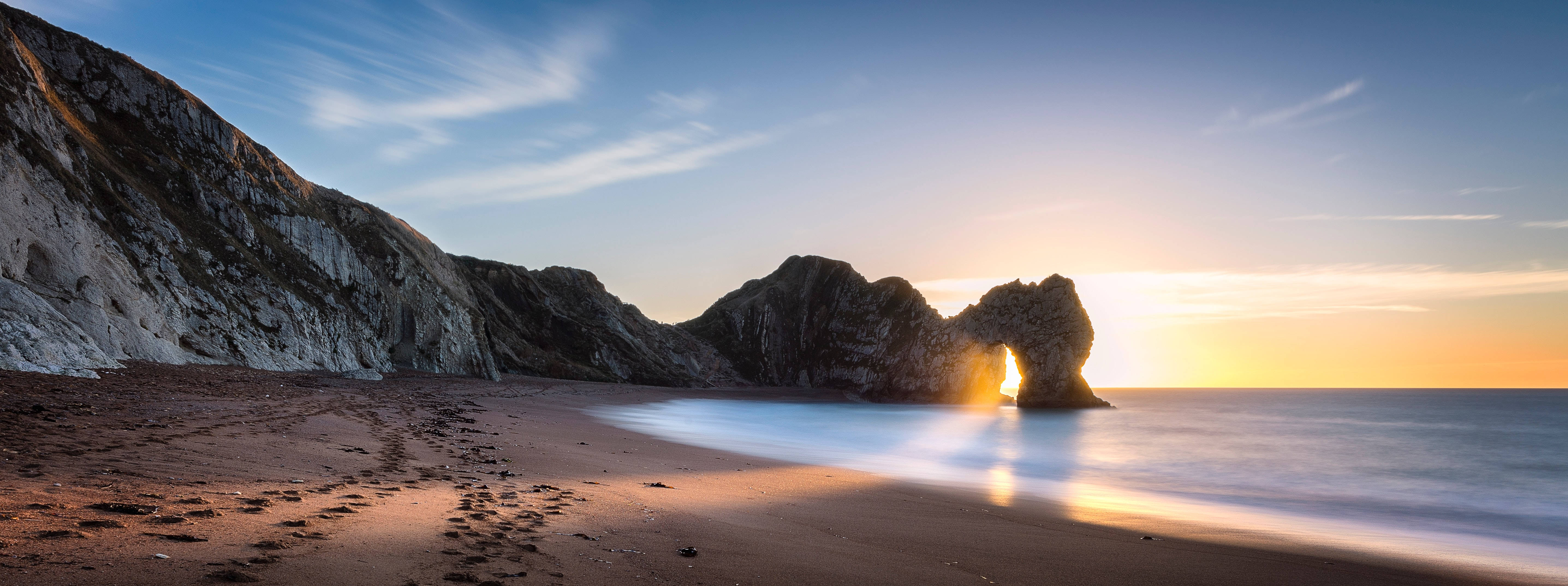 England 桌布 壁紙 — 夕陽穿過標誌性的杜德爾門拱門，在平靜的海面和沙灘上灑下金色的光芒。