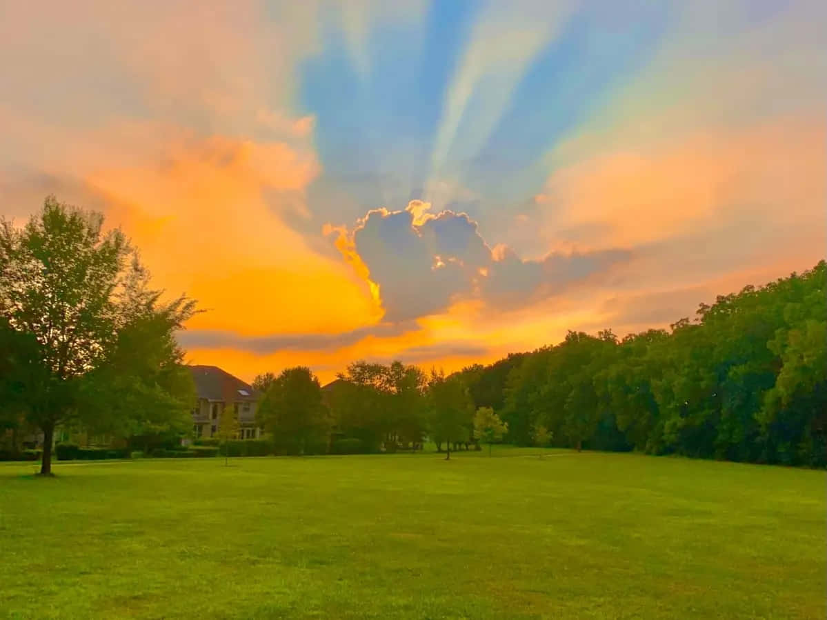 I Love God 桌布 壁紙 — 夕陽穿過雲層，照耀在鬱鬱蔥蔥的綠色公園上，樹木和遠處的房屋沐浴在溫暖的金色光芒中。
