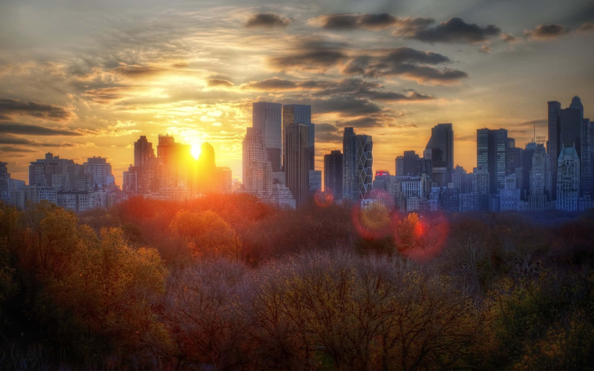 Fall City 桌布 壁紙 — 夕陽穿過雲層照耀在城市天際線上，前景有秋天的樹木。