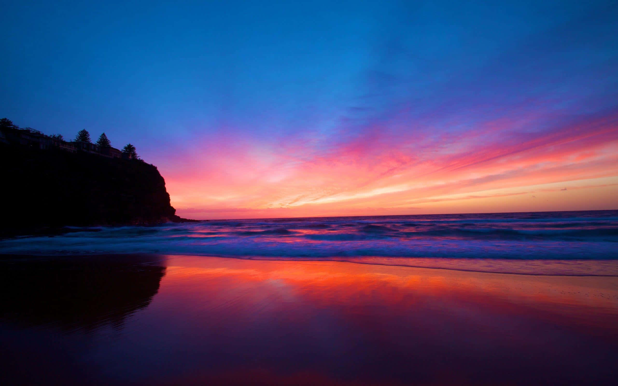 Sunset Landscape 桌布 壁紙 — 夕陽色彩斑斕，將天空染成火橙色和深紫色，海浪拍打著黑暗懸崖旁的濕沙灘。