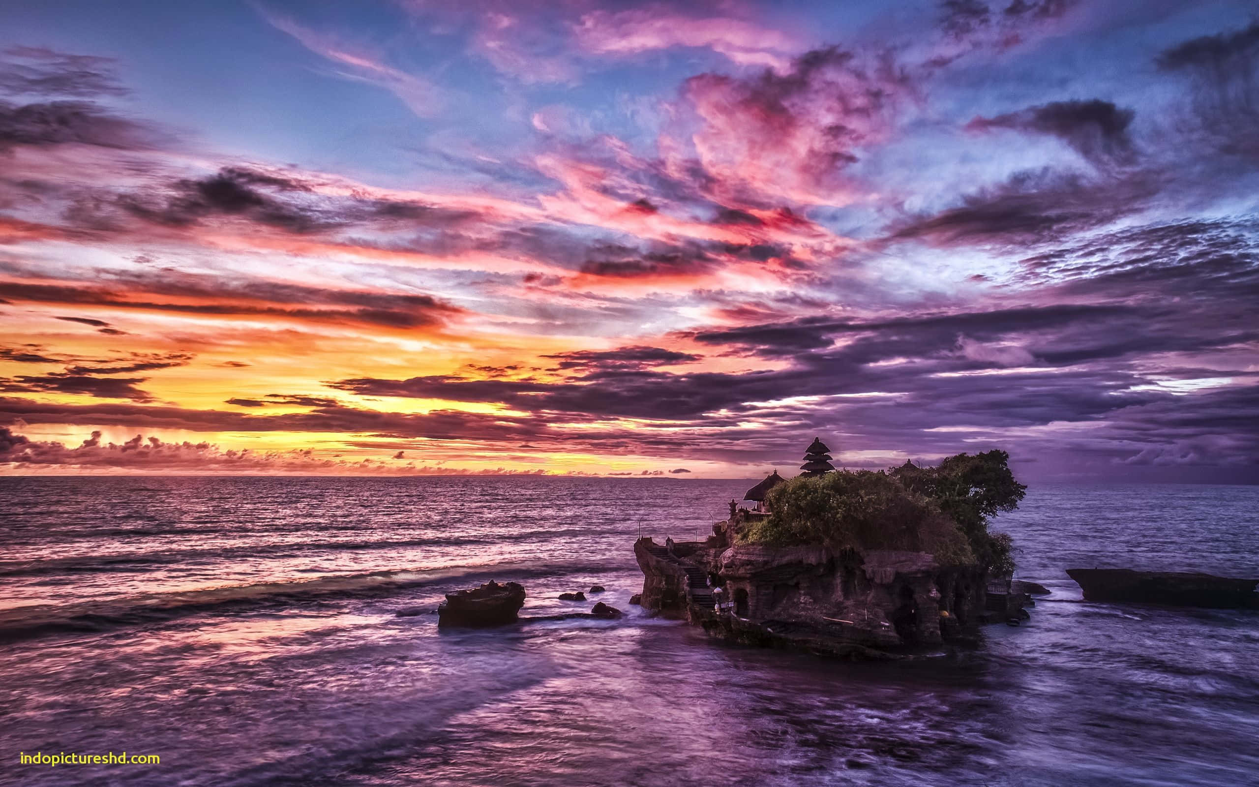 Bali Beach 桌布 壁紙 — 夕陽西下，絢麗的色彩將天空染成火熱的橙色和深紫色，映照著海洋中岩石島上的寺廟。