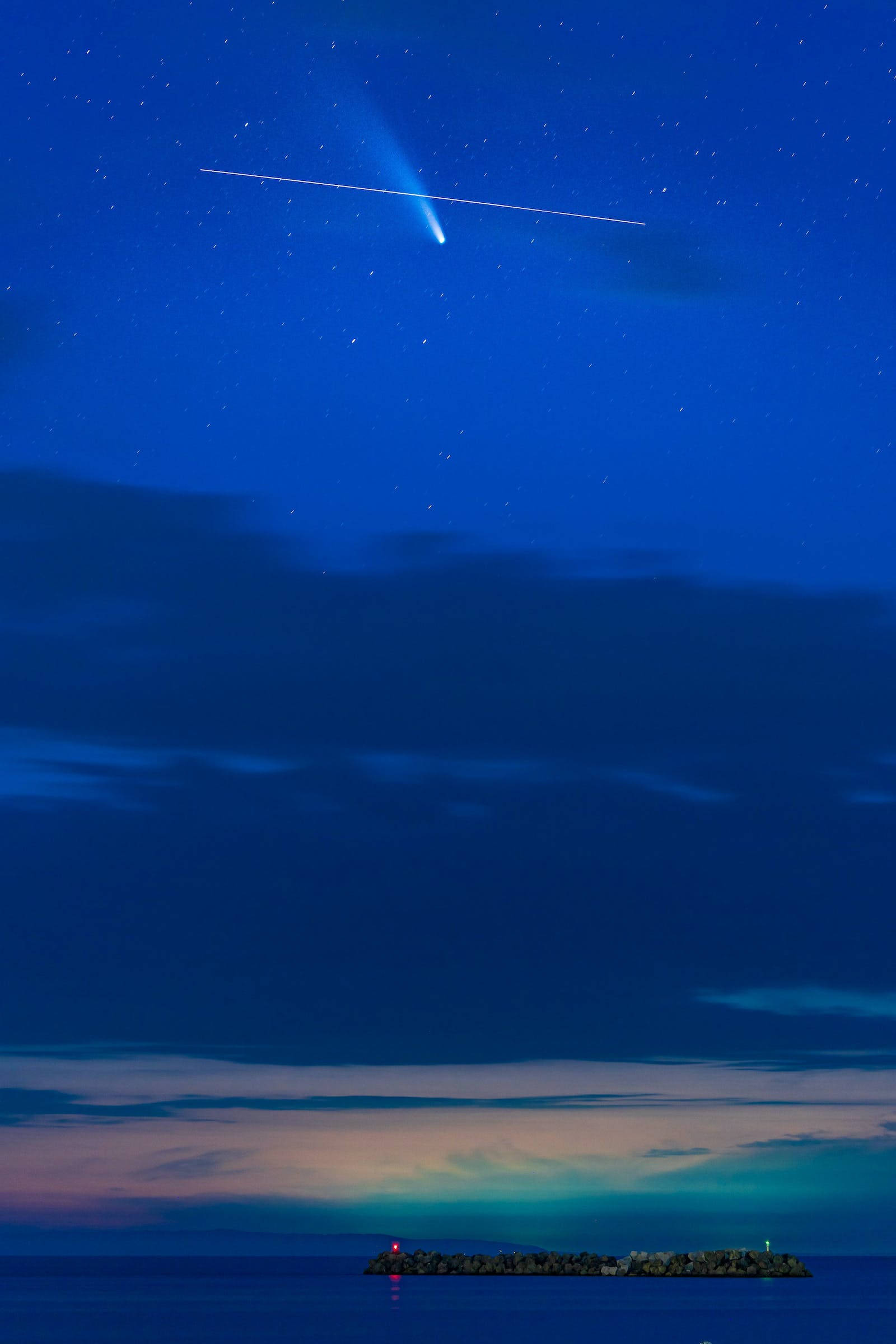 外太空中的藍色星系壯麗景象 壁紙 — 一顆明亮的彗星劃過平靜海面上方的星空夜空，遠處有岩石島嶼和燈塔。