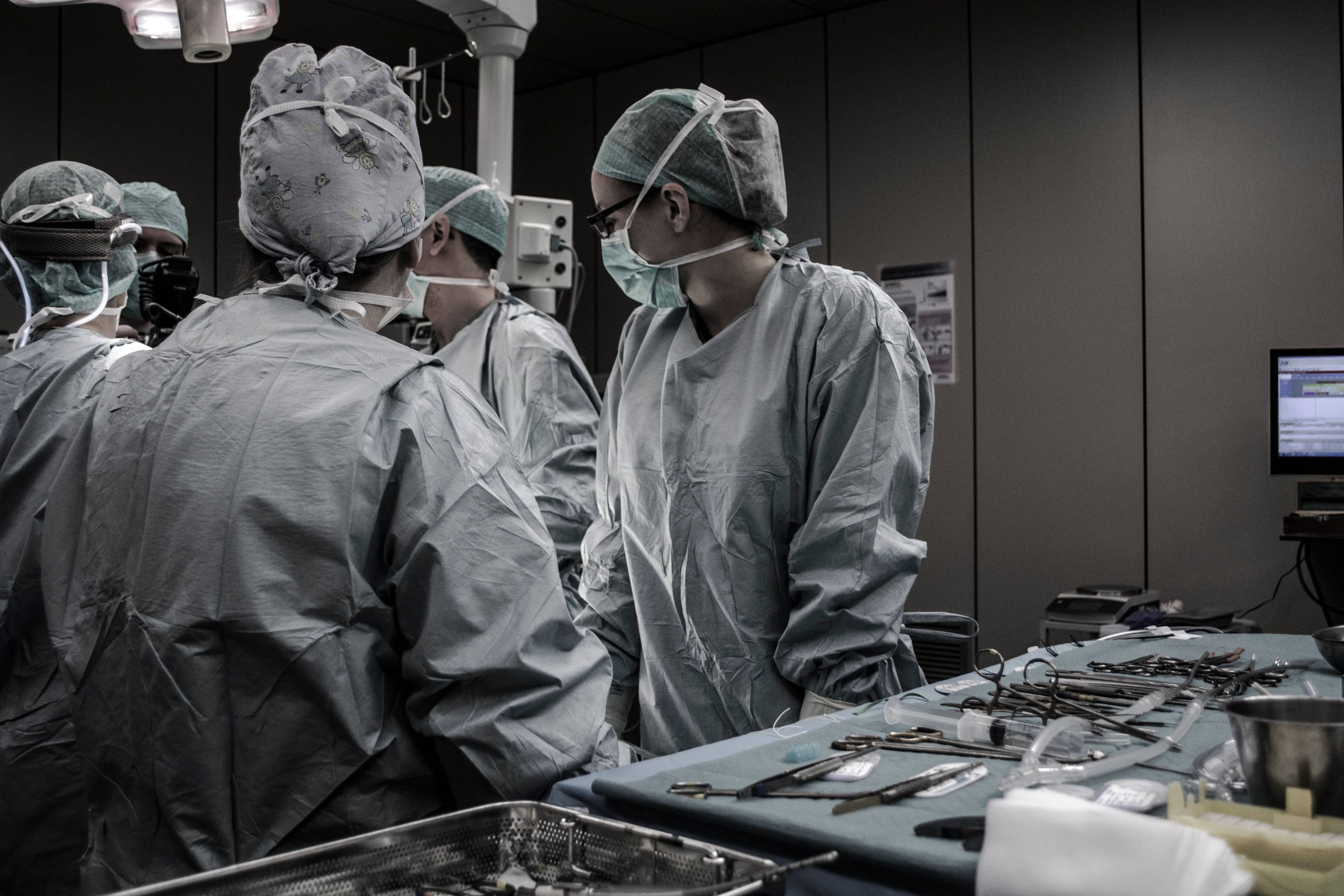 Surgery 桌布 壁紙 — 外科醫生穿著無菌手術袍和口罩，在明亮的頭頂燈光下圍著一張佈滿手術工具的手術枱。