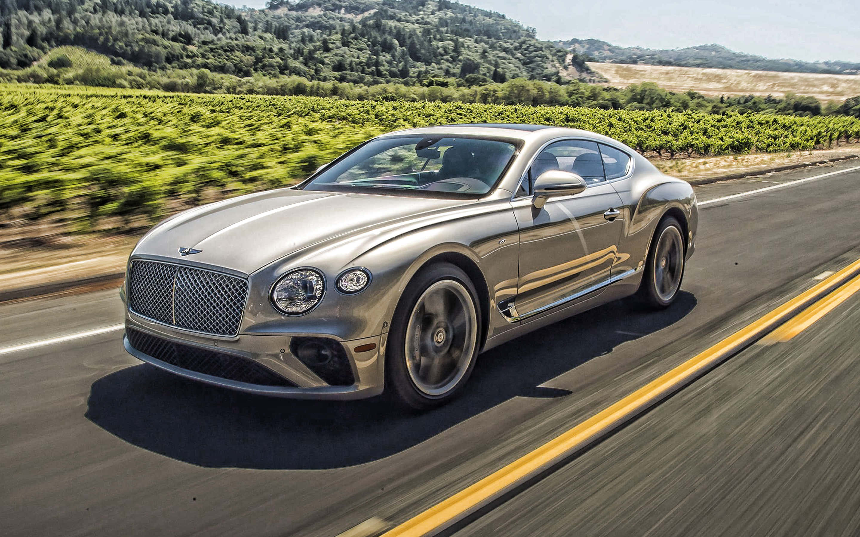 外觀時尚、性能強勁 — 賓利運動車 壁紙 — 一輛銀色賓利 Continental GT 在陽光明媚的道路上行駛，兩旁是鬱鬱蔥蔥的綠色葡萄園和起伏的丘陵。
