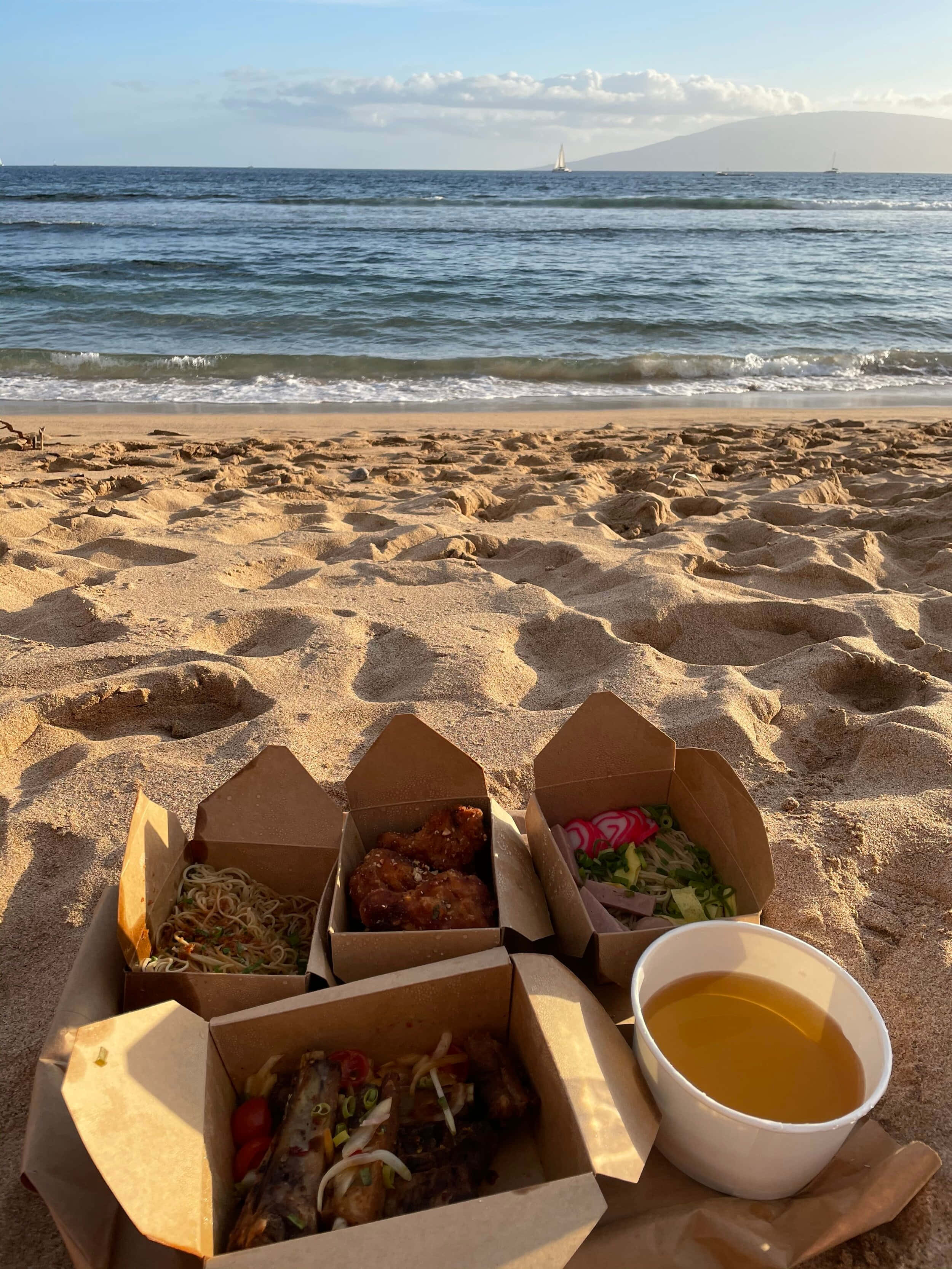 Beach Picnic 桌布 壁紙 — 外賣食物盒和飲品放在沙灘上，背景是海浪和遠處的帆船，天空晴朗。