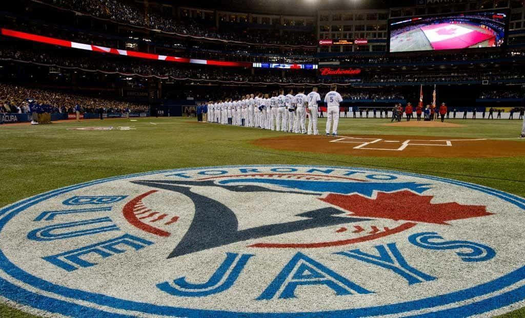 Blue Jays. 桌布 壁紙 — 多倫多藍鳥棒球隊在夜間球場燈光下列隊，草地上有球隊標誌。