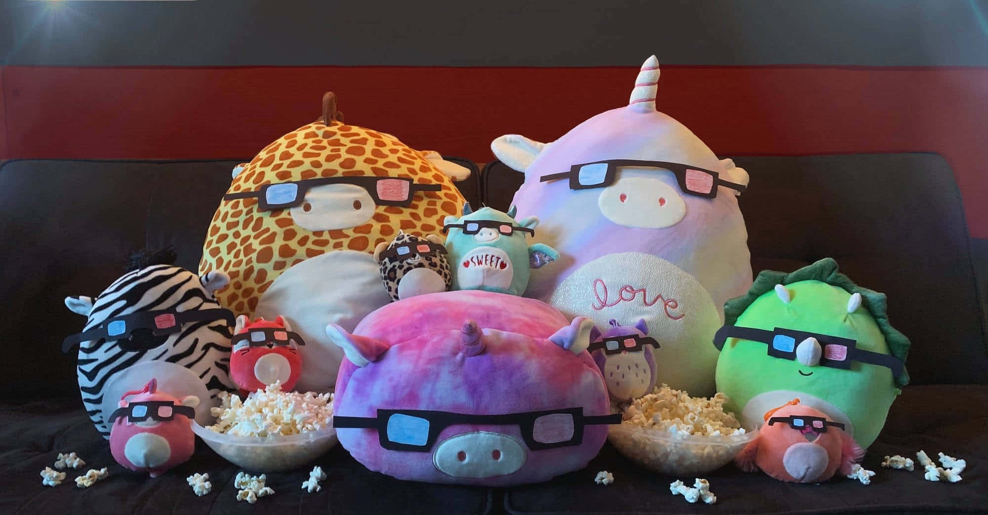 多彩可愛的Squishmallow玩偶集合 壁紙 — 一群色彩繽紛的毛絨動物戴著3D眼鏡和爆谷碗，舒適地坐在深色梳化上。