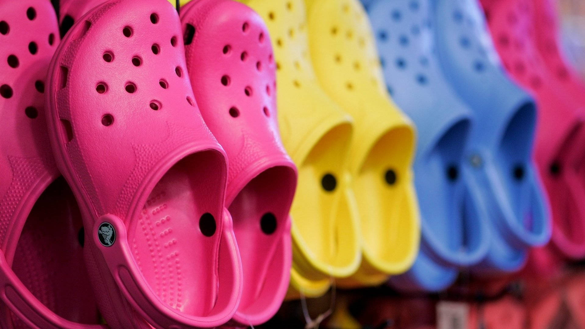 多款Crocs鞋展示 壁紙 — 充滿活力的粉紅色、黃色和藍色Crocs鞋整齊排列在零售展示中，景深較淺。