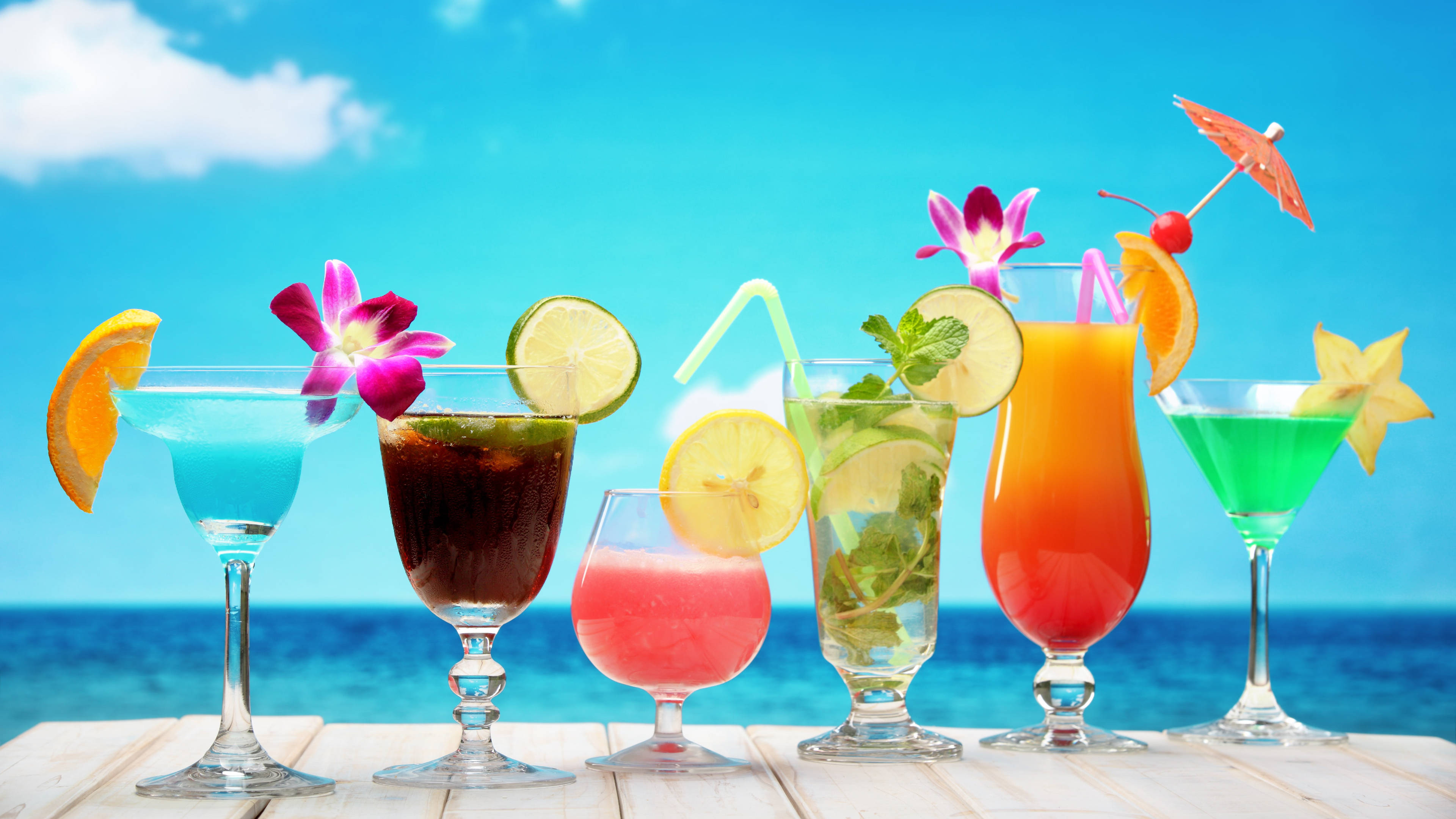 多款熱帶飲品 壁紙 — 色彩繽紛的熱帶雞尾酒配上水果裝飾和鮮花，放在木桌上，背景是明亮的藍色海洋和天空。