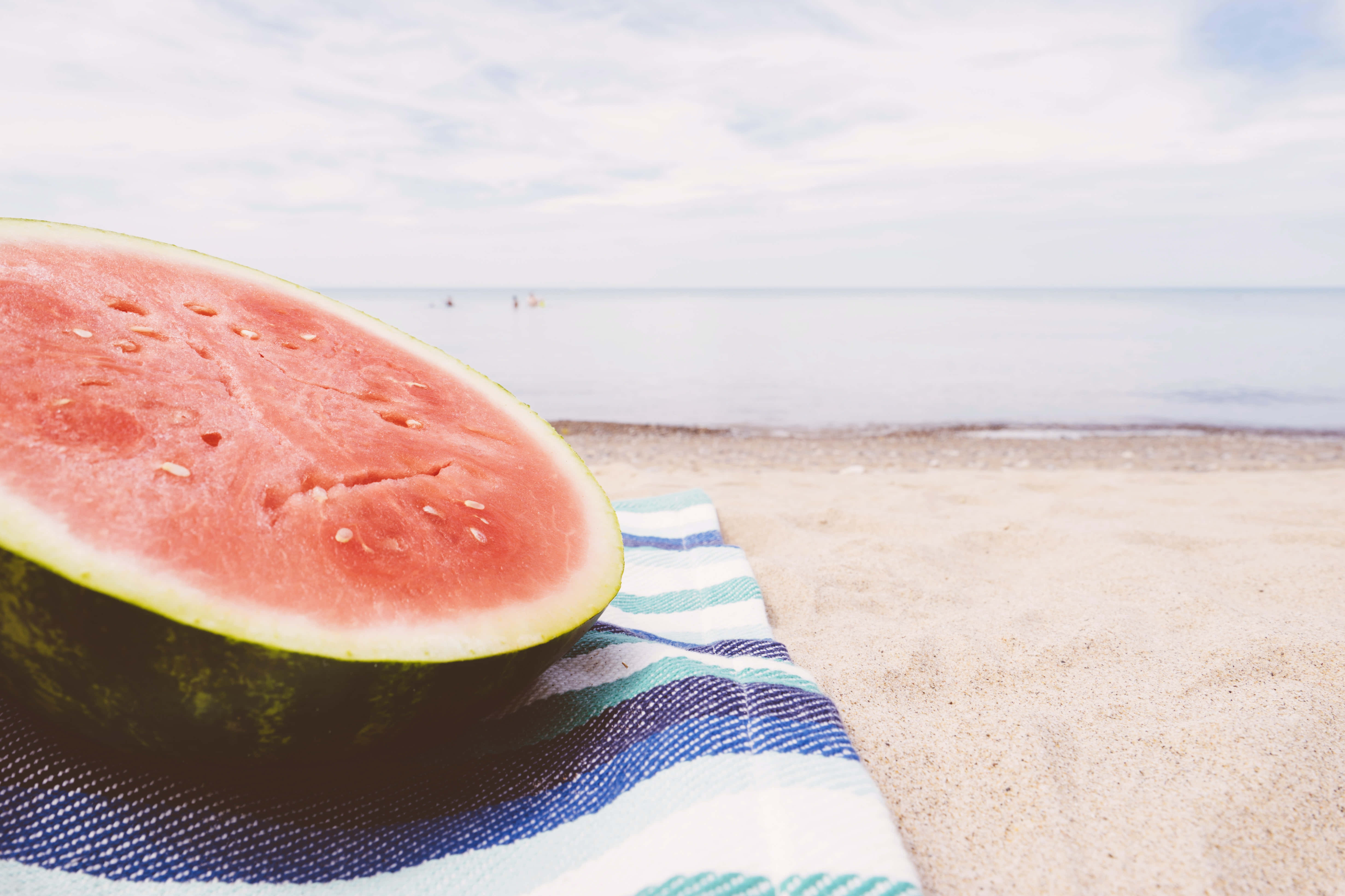 Summer Fruits 桌布 壁紙 — 多汁嘅西瓜半個放喺沙灘上嘅條紋毛巾上面，背景係平靜嘅海洋同多雲嘅天空，柔和焦點。