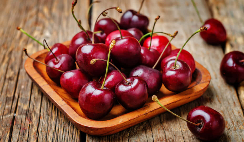 Red Fruit 桌布 壁紙 — 多汁成熟的車厘子連莖在粗糙木碟上閃閃發光，置於經風化的木桌上。