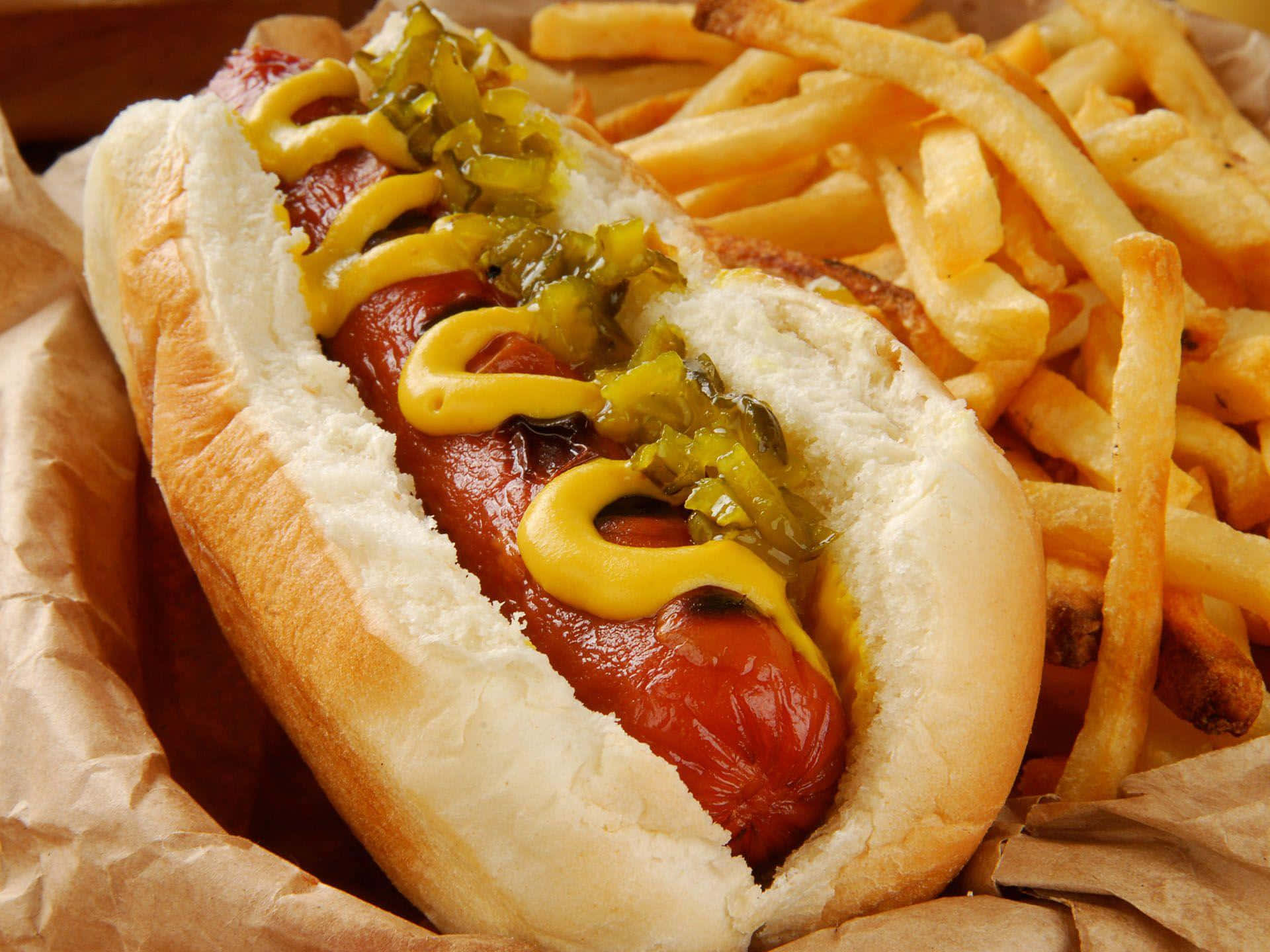 Hot Dog 桌布 壁紙 — 多汁的熱狗配上芥末和調味醬，放在皺巴巴的棕色紙上，旁邊是酥脆的金黃薯條。