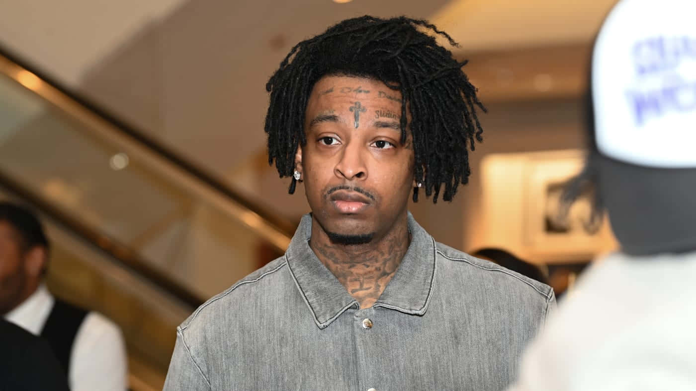 多白金唱片銷量藝人 21 Savage 壁紙 — 一名留著辮子頭、臉上有紋身的男子，在溫暖照明的室內環境中，身後有自動扶梯，他專注地凝視著。