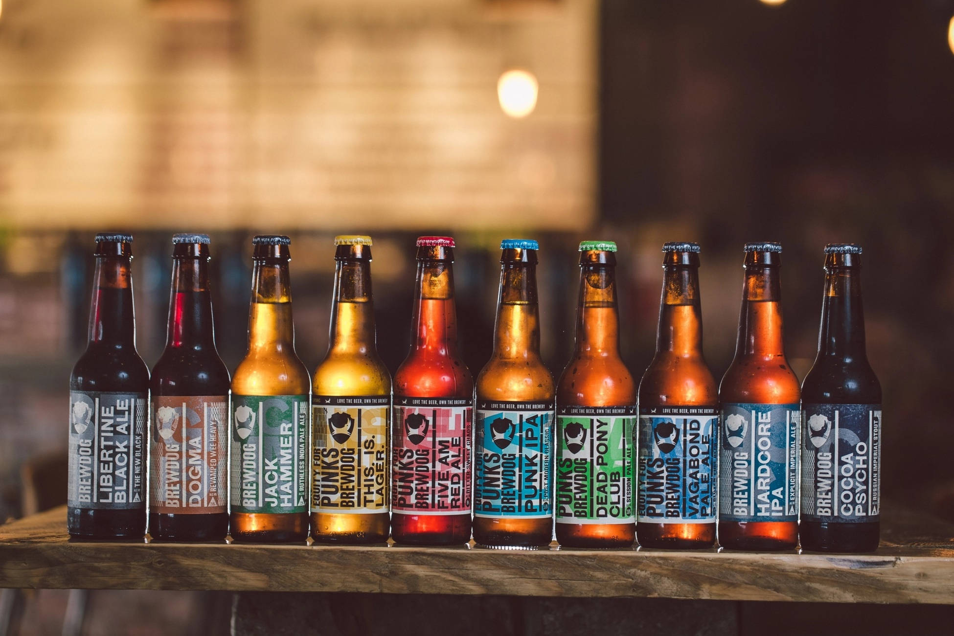 多種 BrewDog 精釀啤酒瓶 壁紙 — 一排色彩繽紛的BrewDog啤酒瓶排列在質樸的木吧上，在溫暖的環境燈光下。