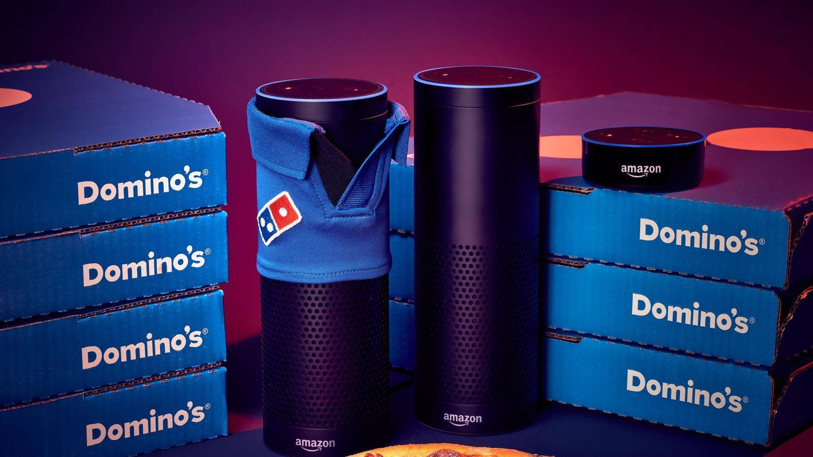 多米諾披薩商品 壁紙 — Amazon Alexa 裝置藏在堆疊的 Domino's 薄餅盒之間，在充滿活力的紫色燈光下。