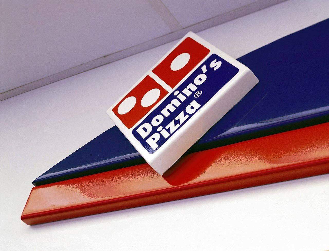 多米諾披薩斜角標誌 壁紙 — 特寫鏡頭：一塊 Domino's Pizza 標誌牌擱在紅色和藍色傾斜面板上，背景為白色天花板。