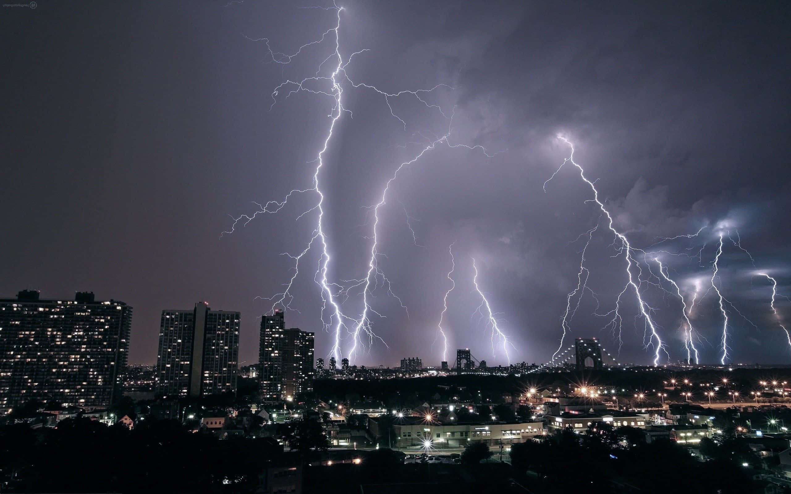 thunder 桌布 壁紙 — 多道鋸齒狀閃電照亮了暴風雨夜空中帶有發光窗戶和遠處橋樑的城市景觀。